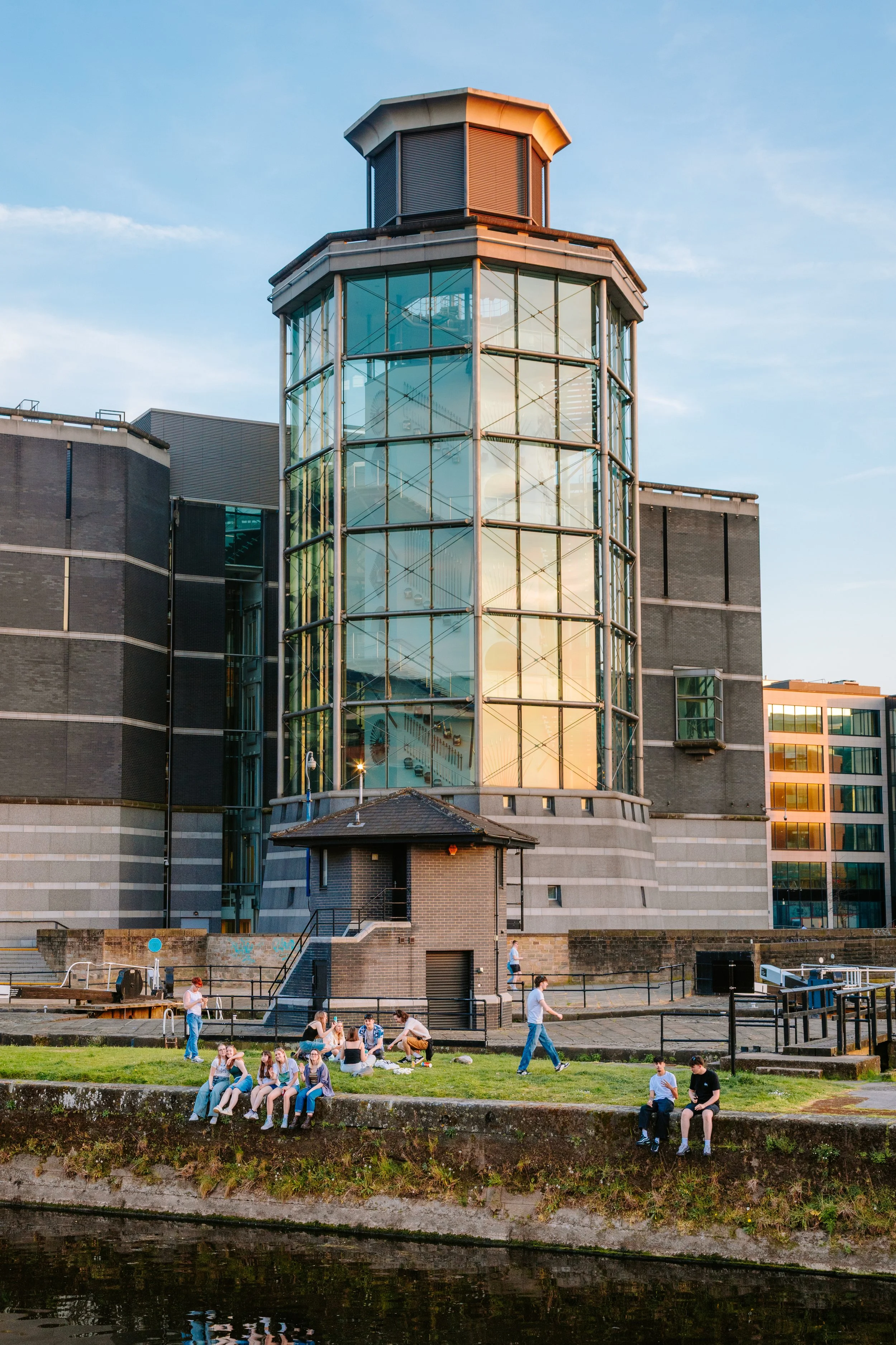 People sitting and standing along a riverbank in front of a modern building with glass windows and a tower, during sunset. Royal armouries leeds docks leeds city centre.