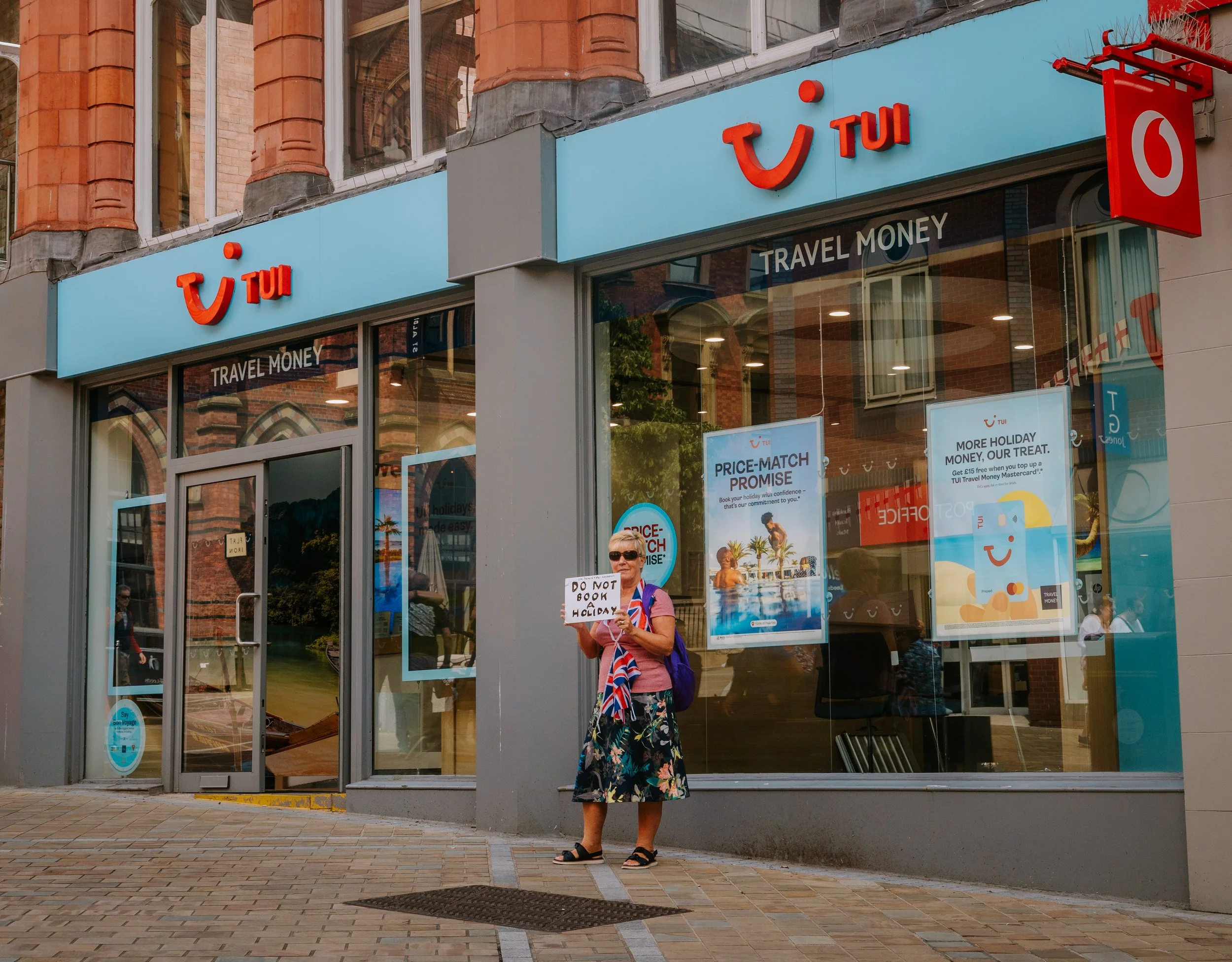 A woman stands outside a TUI travel agency holding a sign that says 'DO NOT BOOK A HOLIDAY.' She is wearing sunglasses, a blue backpack, a pink shirt, a floral skirt, and sandals. The store has large glass windows and promotional posters inside, with