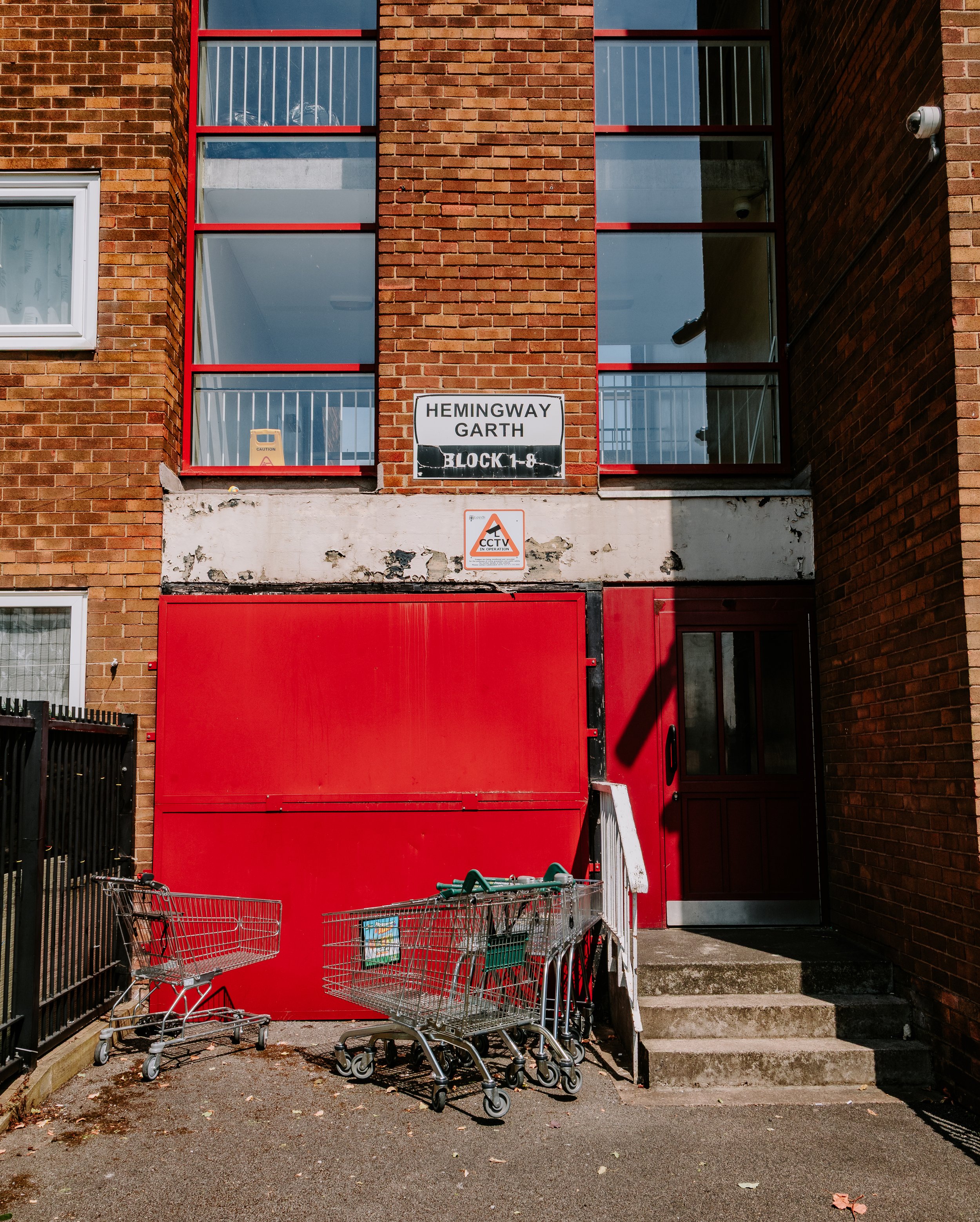 Red fire door with a brick apartment building behind, balconies with glass and metal railing above. A sign reading 'Hemingway Garth Block 1-8' is on the wall. Several shopping carts are parked near the stairs and sidewalk. Hunslet Leeds.