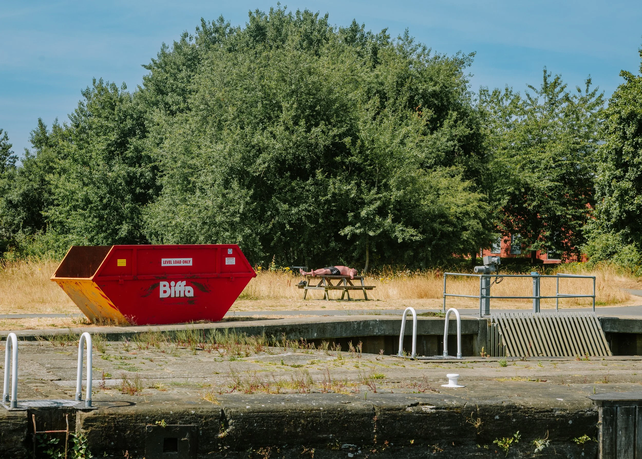 A large green tree in the background, a red skip container with the word "Biffa" and "LEVEL LOAD ONLY" on it, a person lying on a wooden bench, picnic tables, and various metal structures in an outdoor area with grass and pavement.