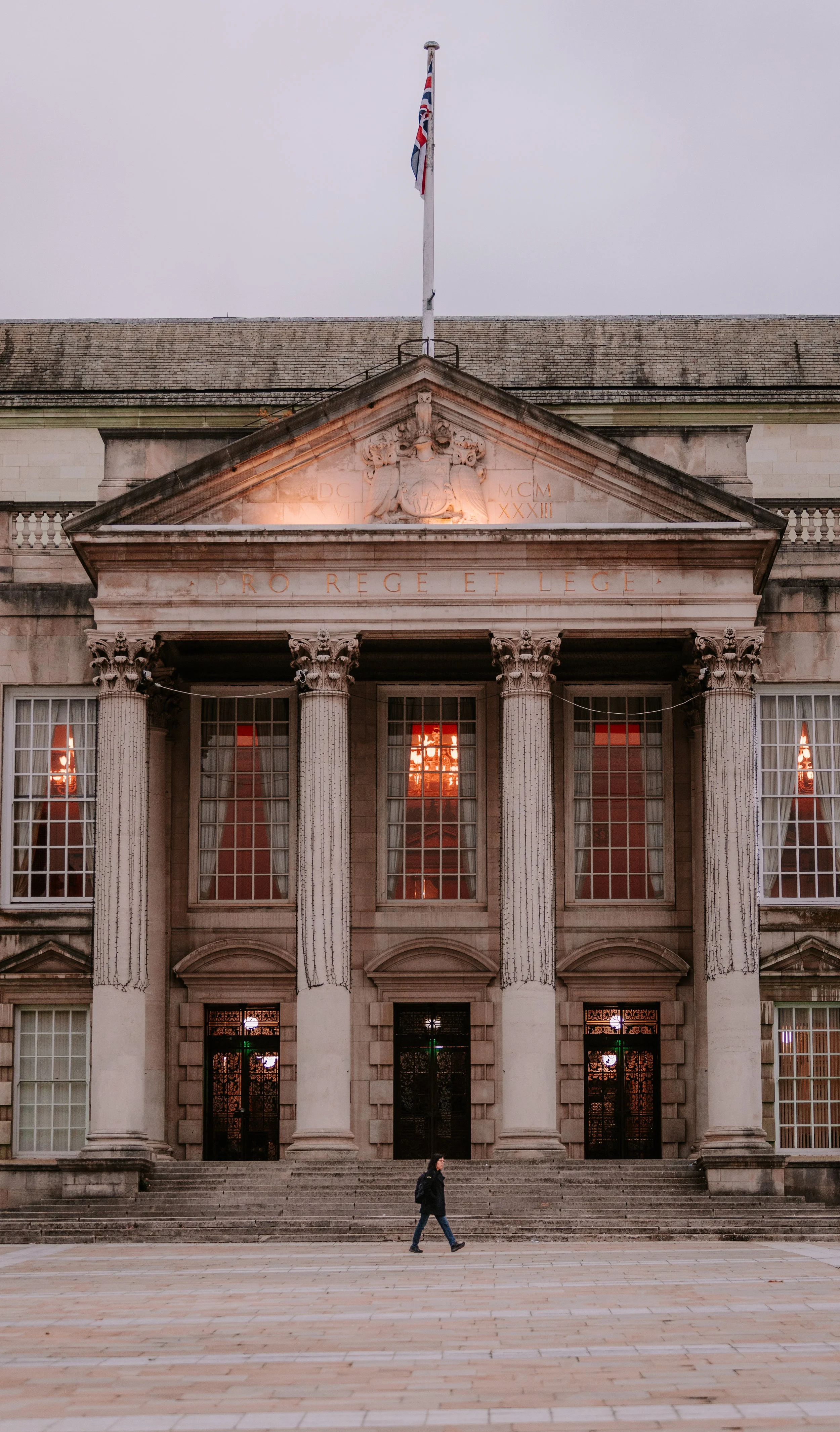 A person walking in front of a historic building with tall columns and a triangular pediment, featuring a flagpole with a Union Jack flag on top and illuminated sculptures and engravings on the facade. leeds city centre millennium square civic hall.