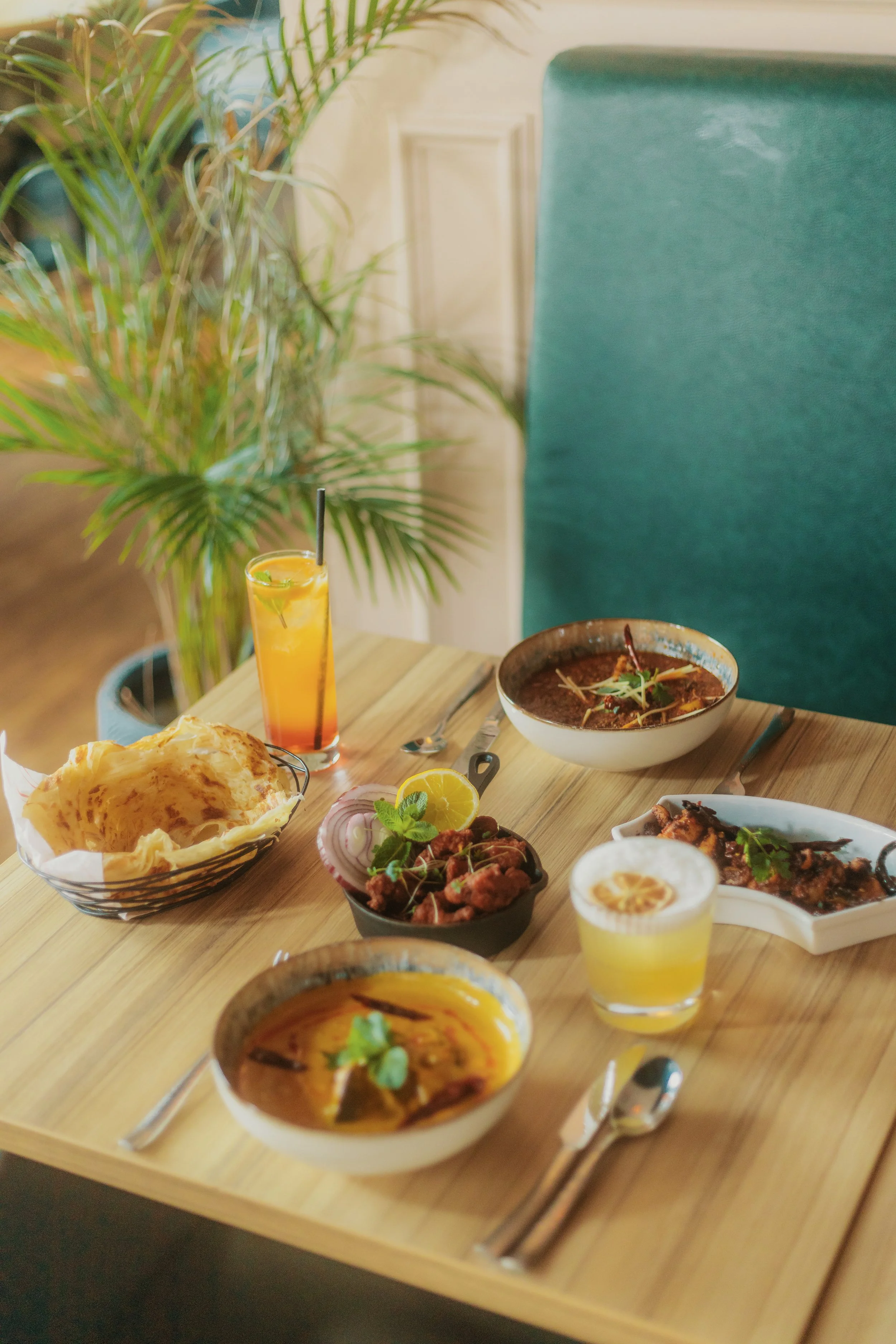 A wooden table with various Indian dishes, a bowl of curry, naan bread, a lemon wedge, and drinks, with a green plant in the background. york leeds yorkshire restaurant photographer videographer