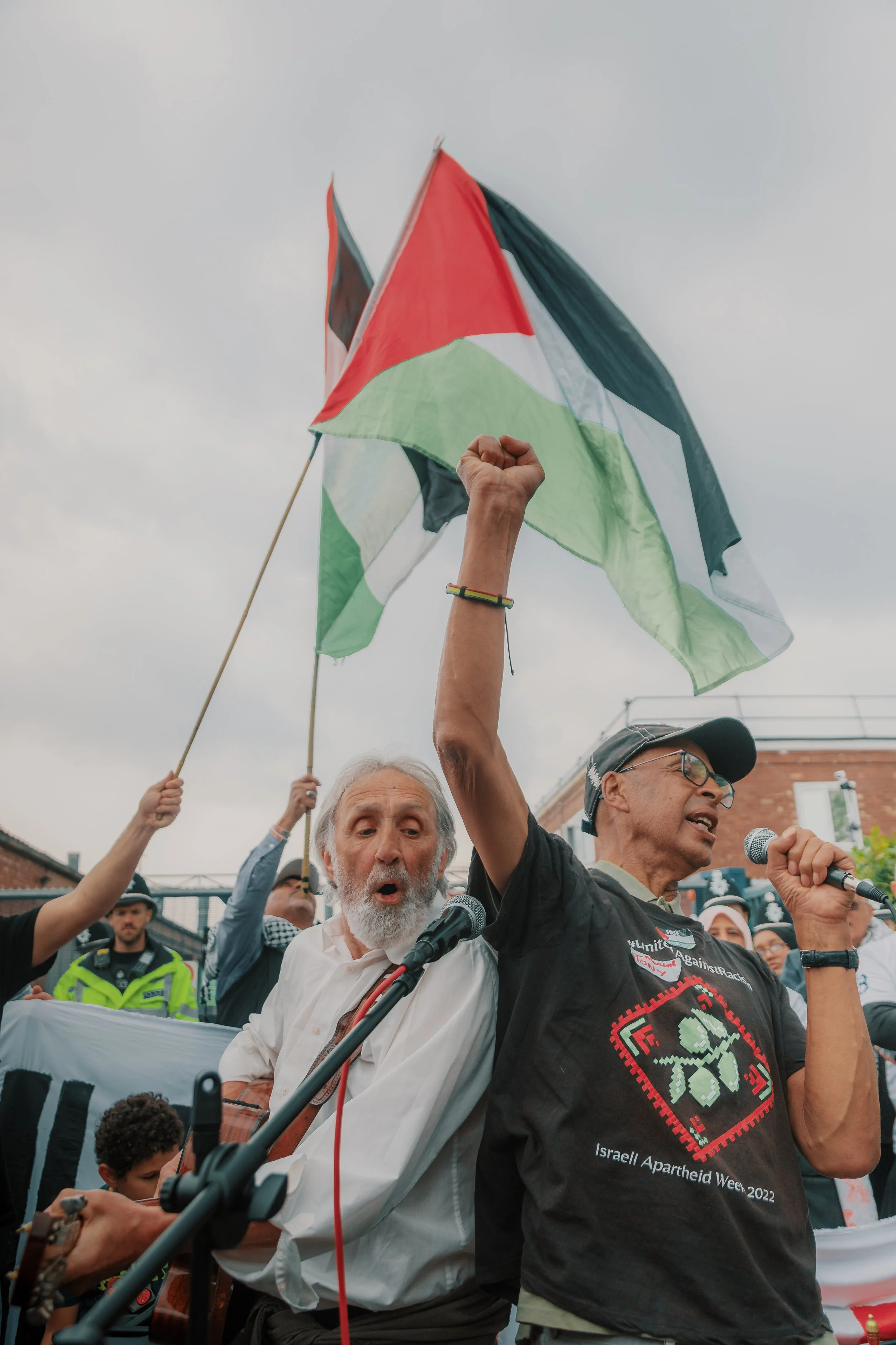 A crowd of protesters, including an older man with a beard and a man holding a Palestinian flag, gathered outdoors. The man with the flag is speaking into a microphone, and there are police officers and other people in the background.