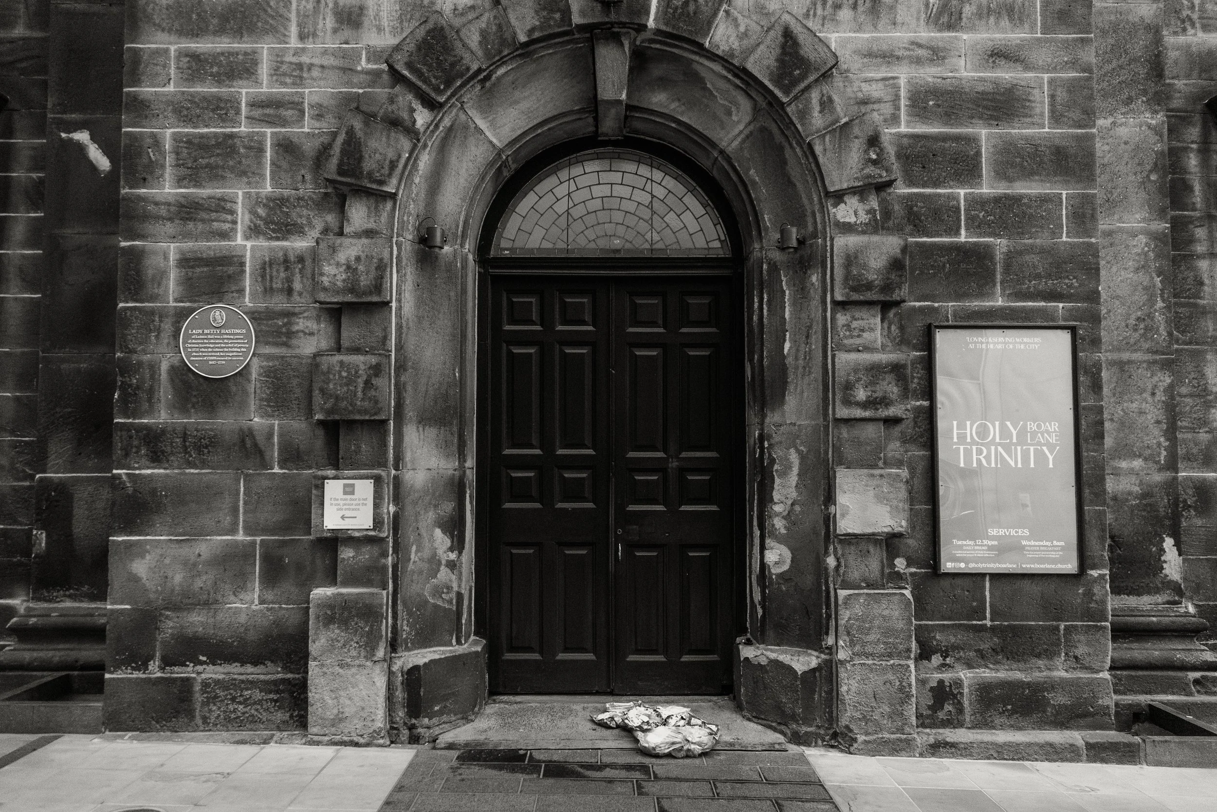 Black and white photo of a stone church entrance with a wooden door and an arch window above it, featuring memorial plaques on either side of the door and a signboard on the right.