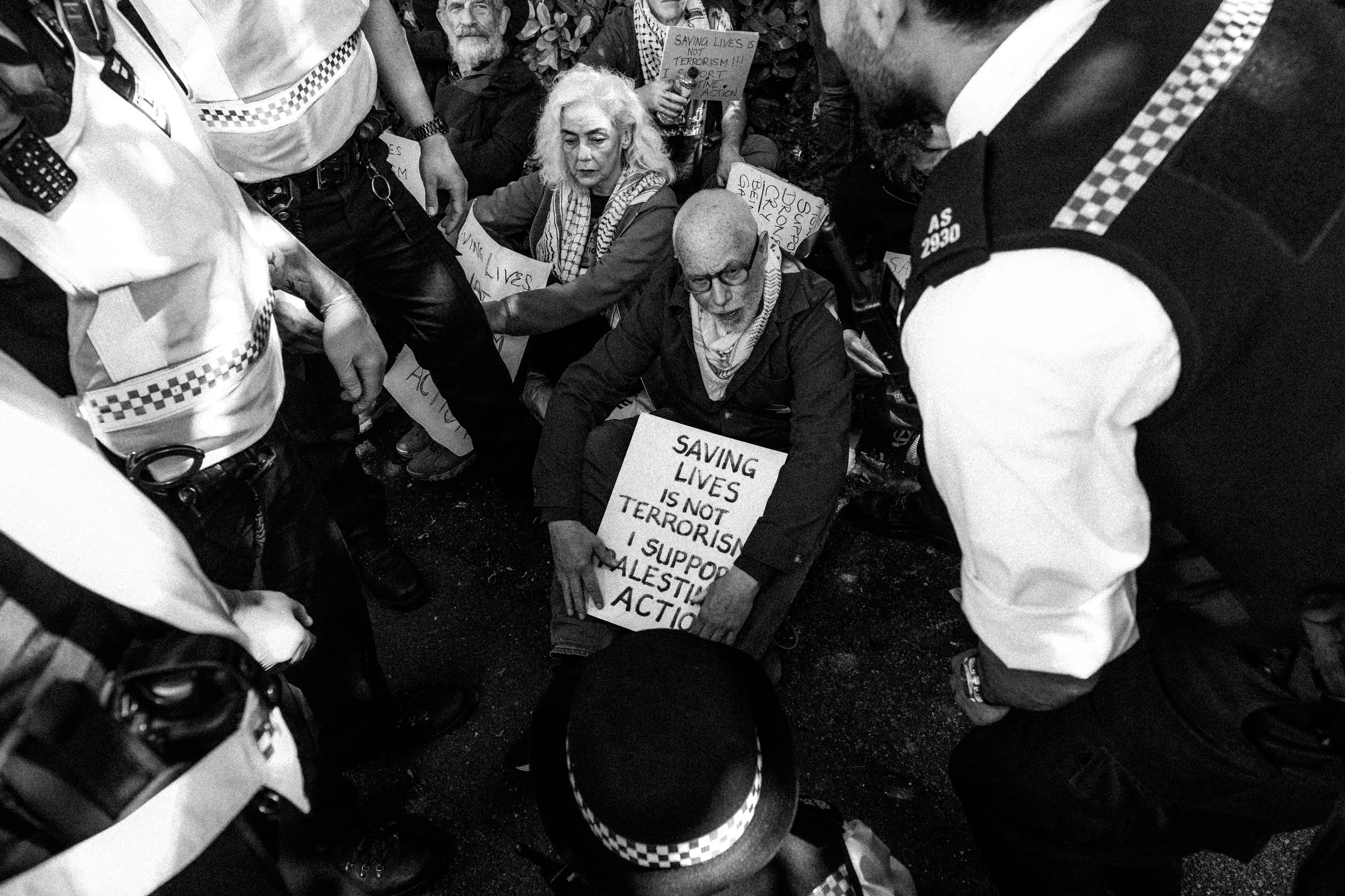 Protesters sitting on the ground holding signs with messages about saving lives and opposing terrorism. Police officers are surrounding them, creating a tense scene.