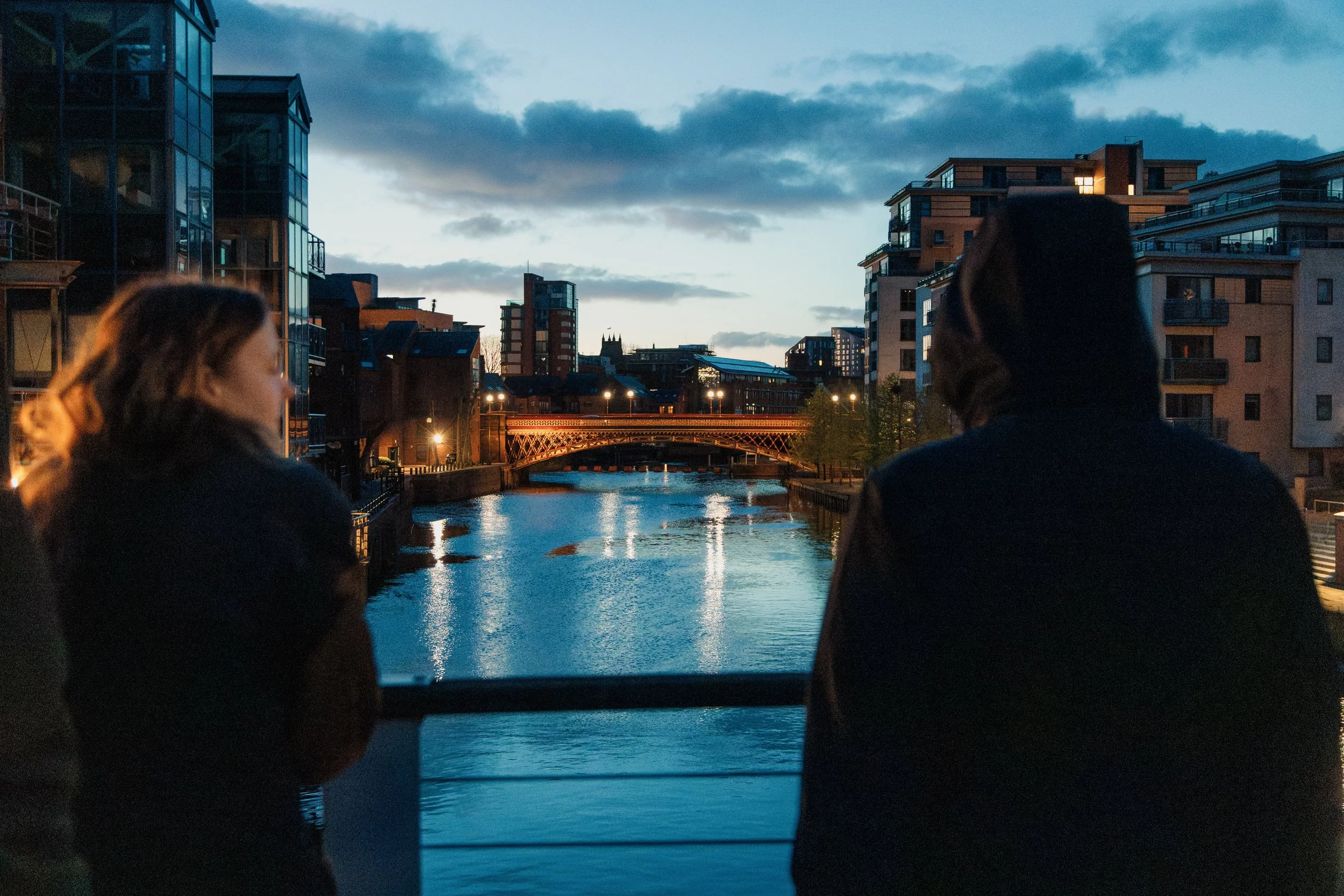 Two people are standing on a bridge over a canal in an urban area during dusk, with modern buildings and cloudy sky in the background.
