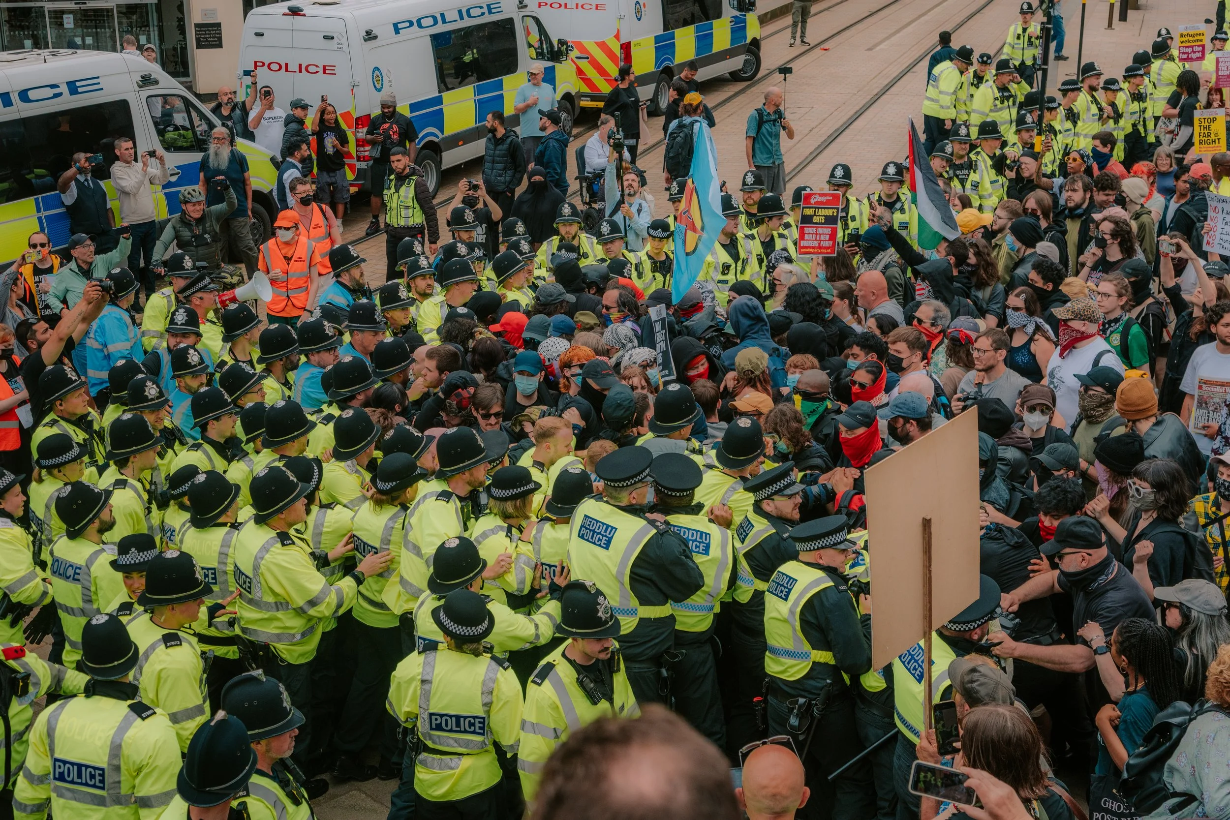 A large crowd of protesters and police officers gathers on a city street during a demonstration. Police officers, wearing yellow reflective vests and black helmets, stand in a line facing the protesters. Several police vehicles are parked behind them
