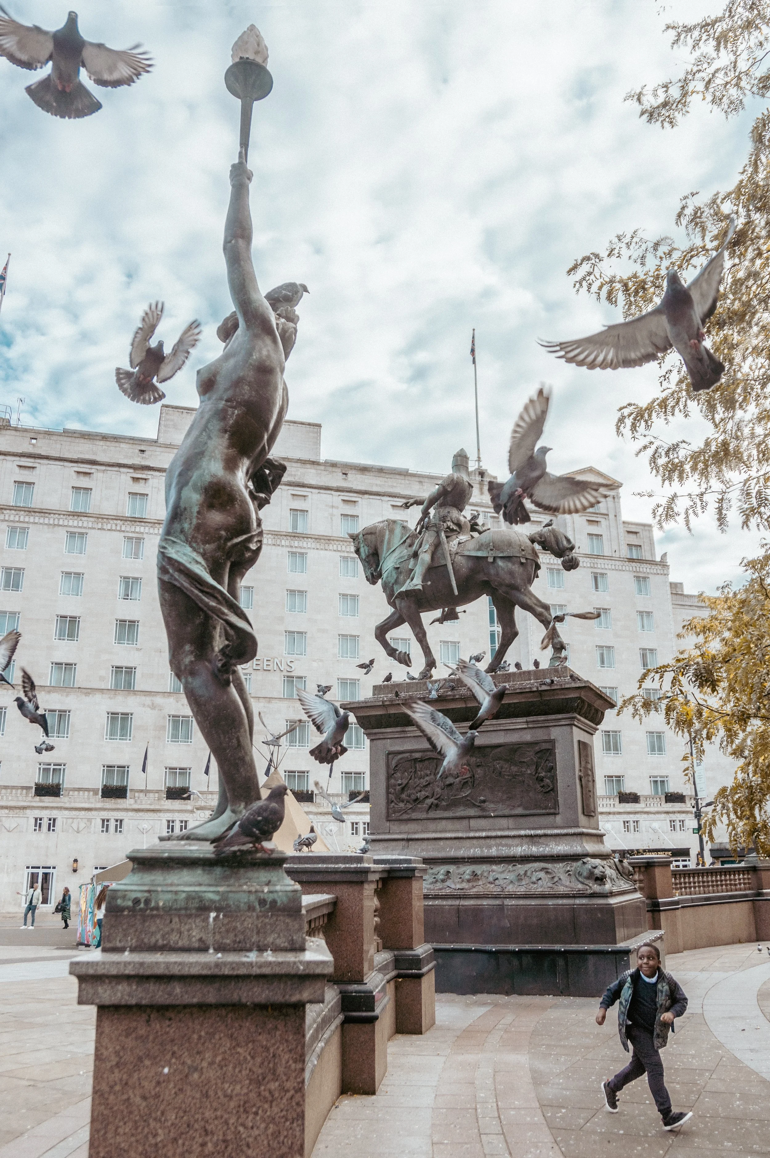 A bronze statue in a city square leeds depicting a person with a bird on their shoulder and several birds flying around. A child is running in the foreground with other people and a large building in the background.