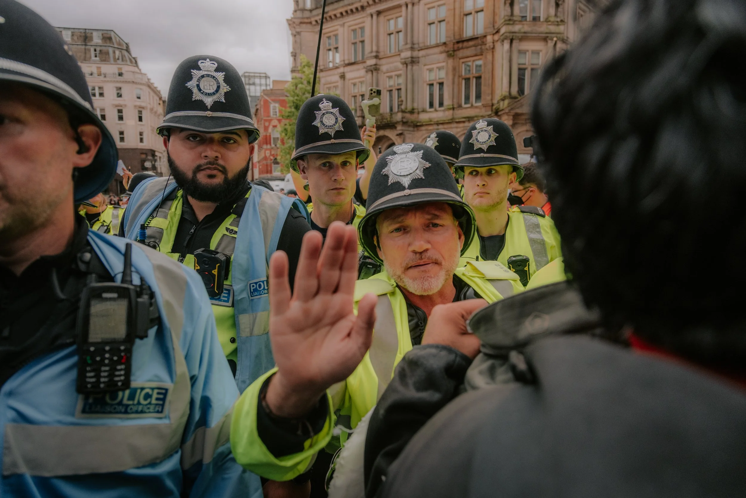 A group of police officers in uniform and helmets facing each other during a confrontation in an Birmingham with historical buildings in the background.
