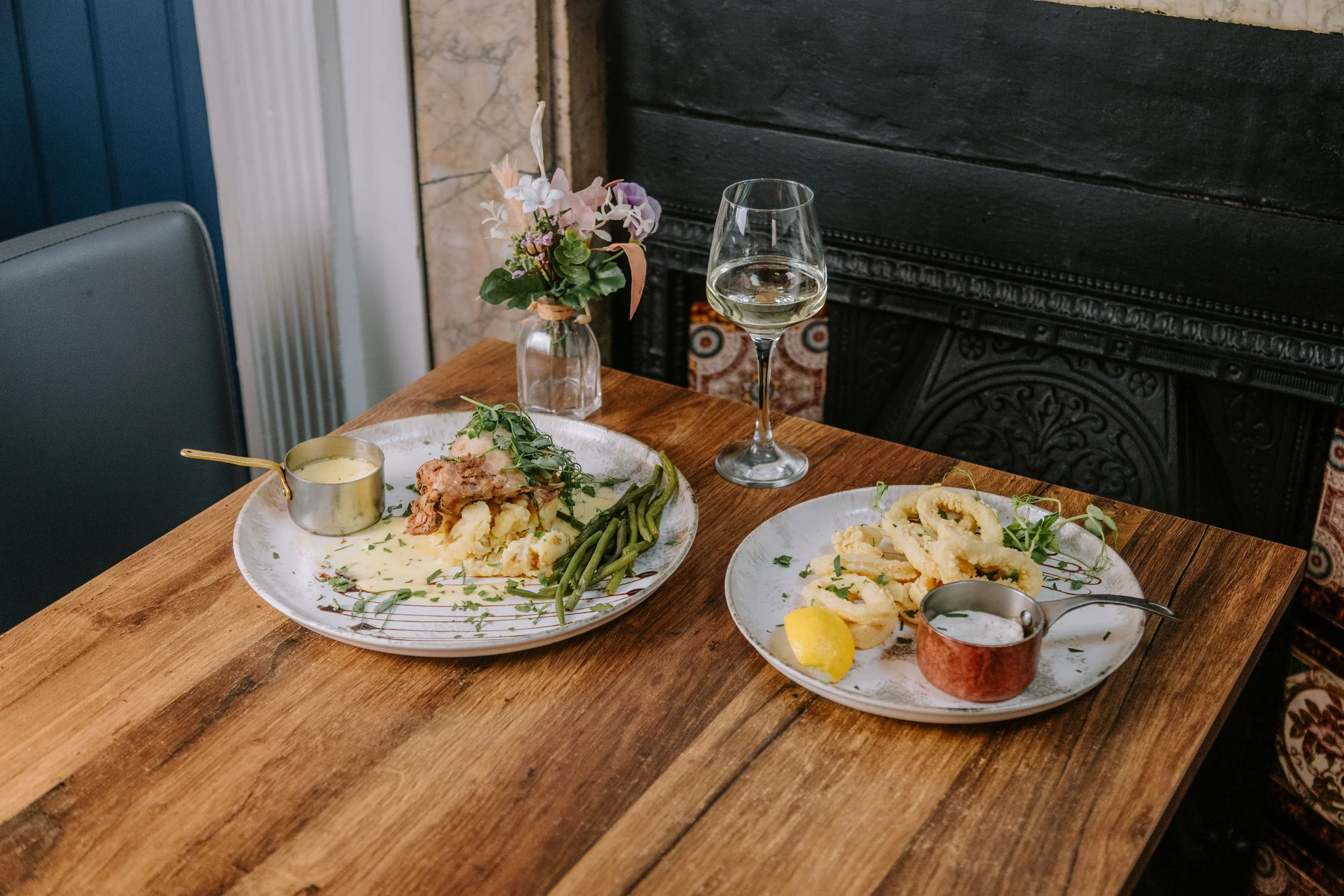 A dining table set with plates of food, including mashed potatoes with meat, green beans, and fried calamari, with a glass of white wine, a small pitcher of sauce, and a vase with flowers in the background. york leeds yorkshire restaurant photography