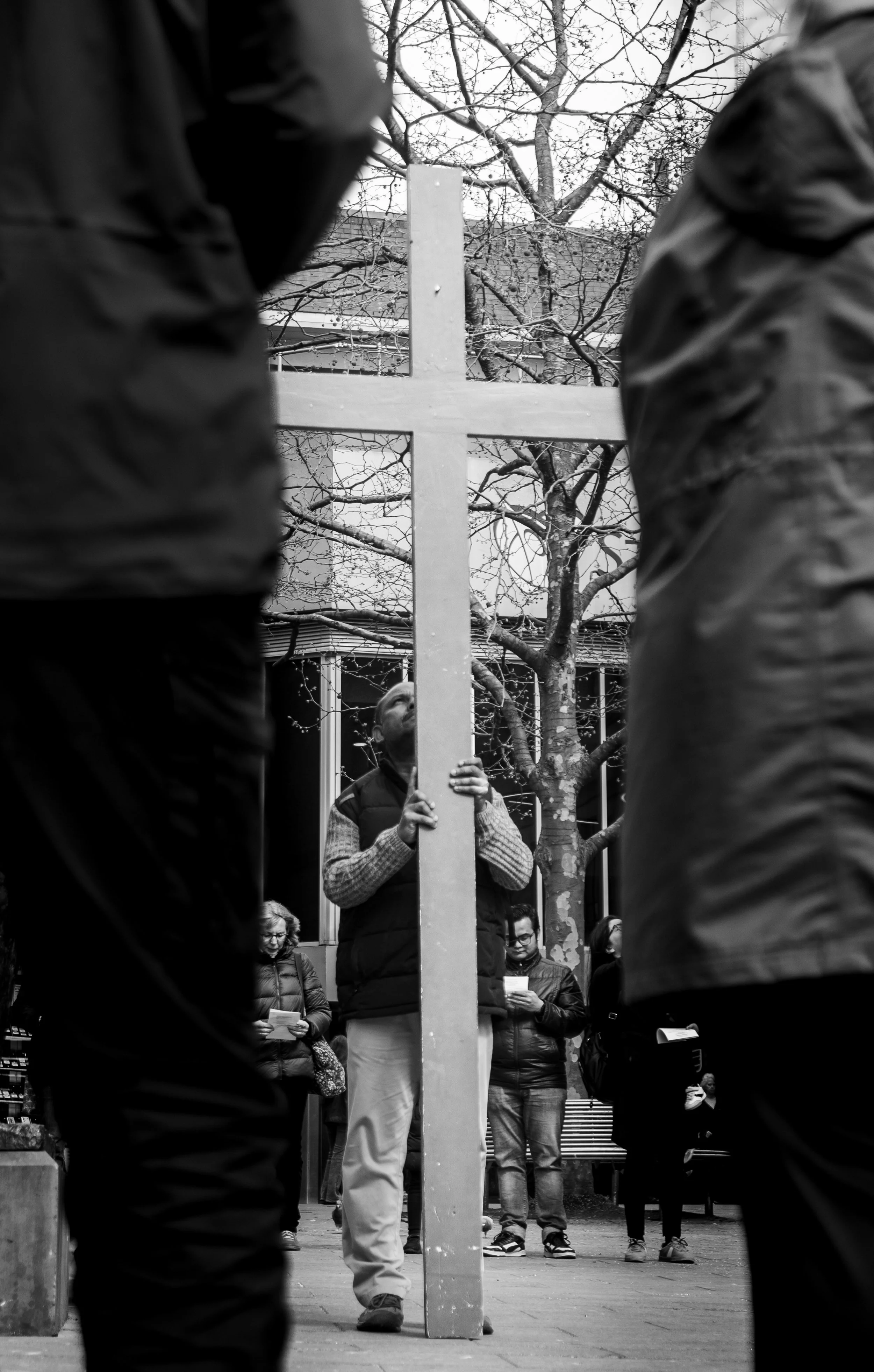 People gathered outdoors around a large cross-shaped object, with some reading papers. The photo is framed by two individuals in the foreground and shows trees and a building in the background.
