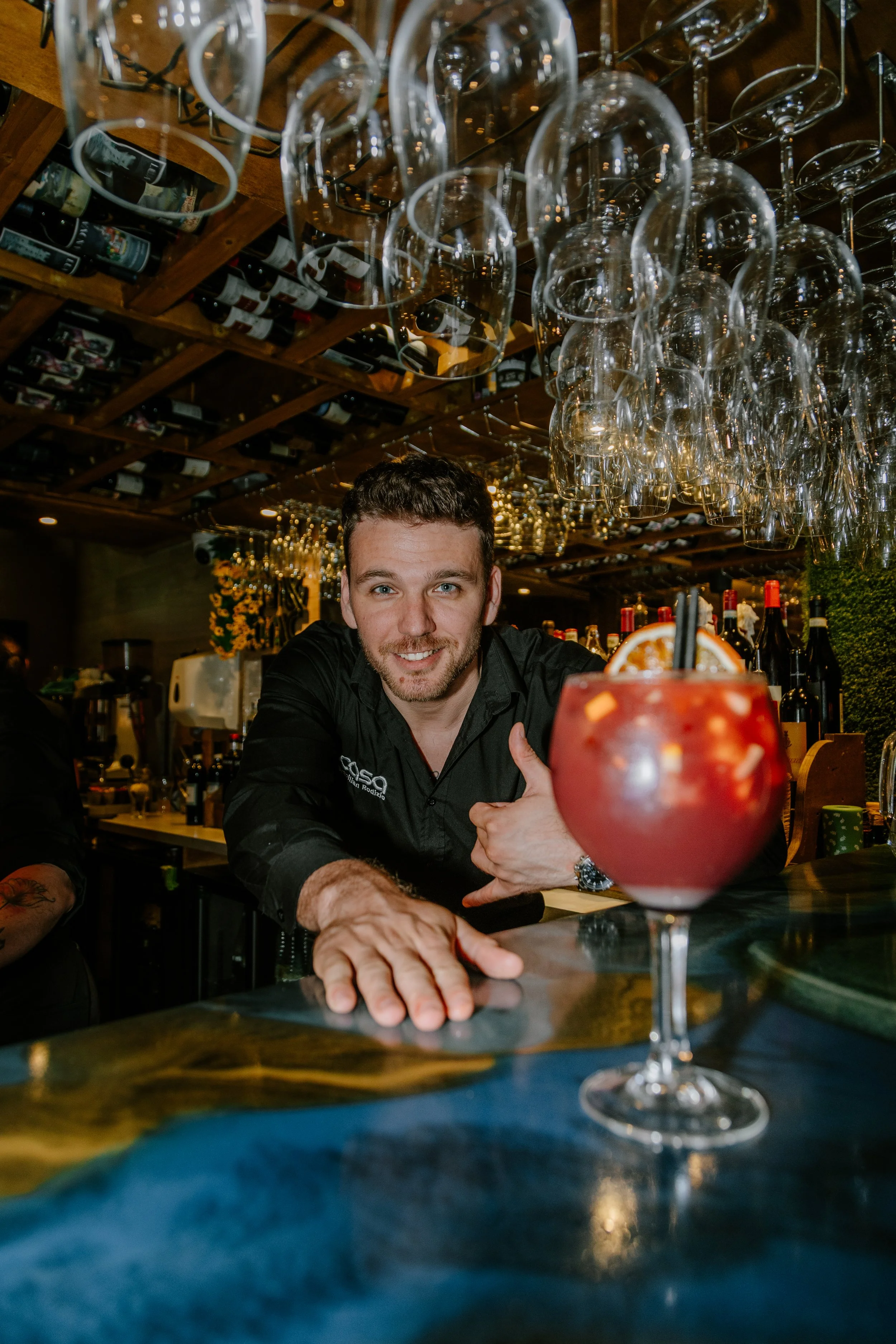 A smiling bartender in a black shirt extends his hand on a bar counter with a red cocktail garnished with citrus and a straw in front of him. Overhead shelves filled with wine bottles and glasses are visible in the background. york leeds yorkshire.