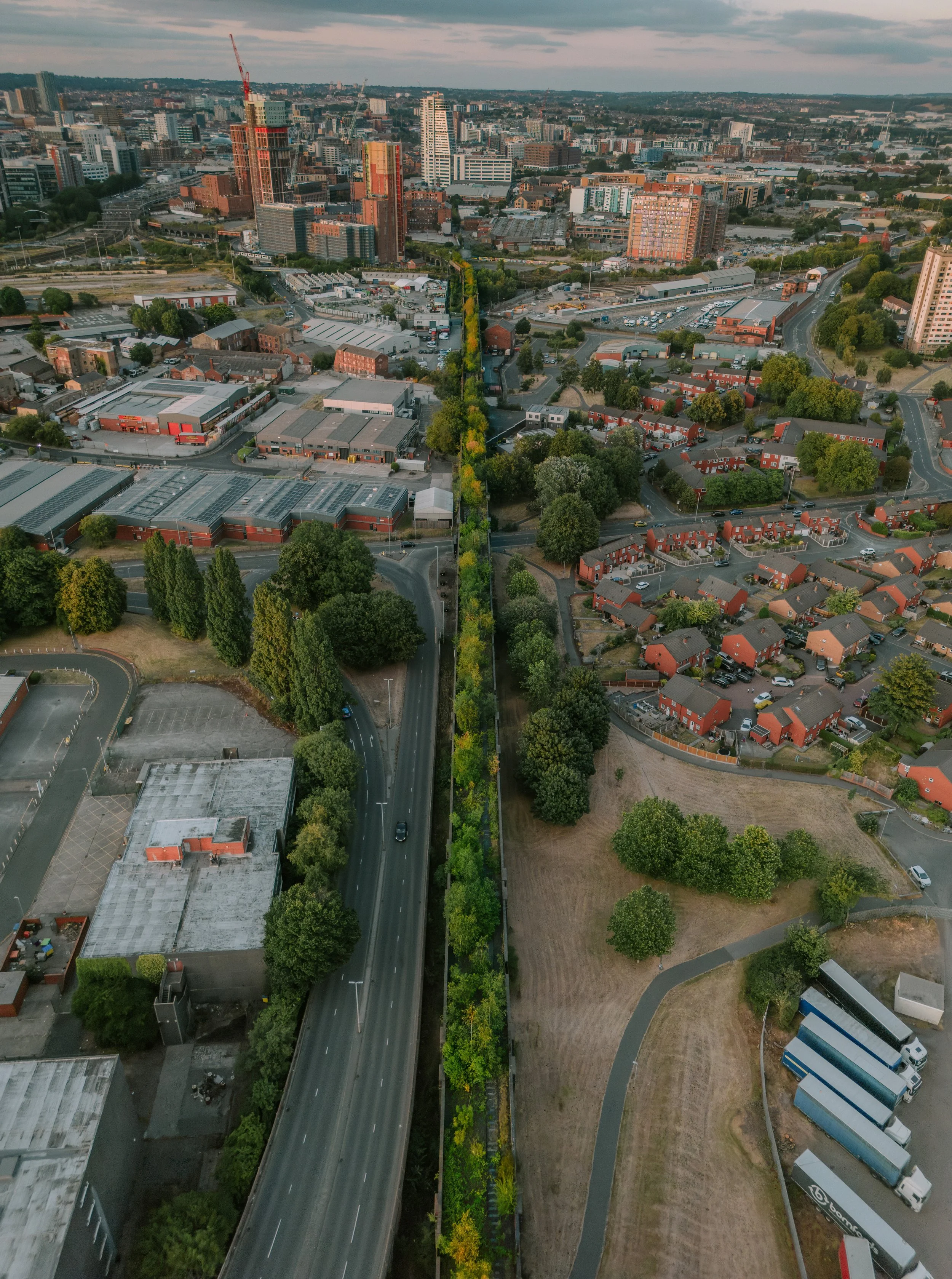 An aerial view of a cityscape with a mix of high-rise buildings, warehouses, residential houses, and green spaces. A main road runs vertically through the image, bordered by trees and various structures. shoiwng holbeck viaduct in leeds south leeds 