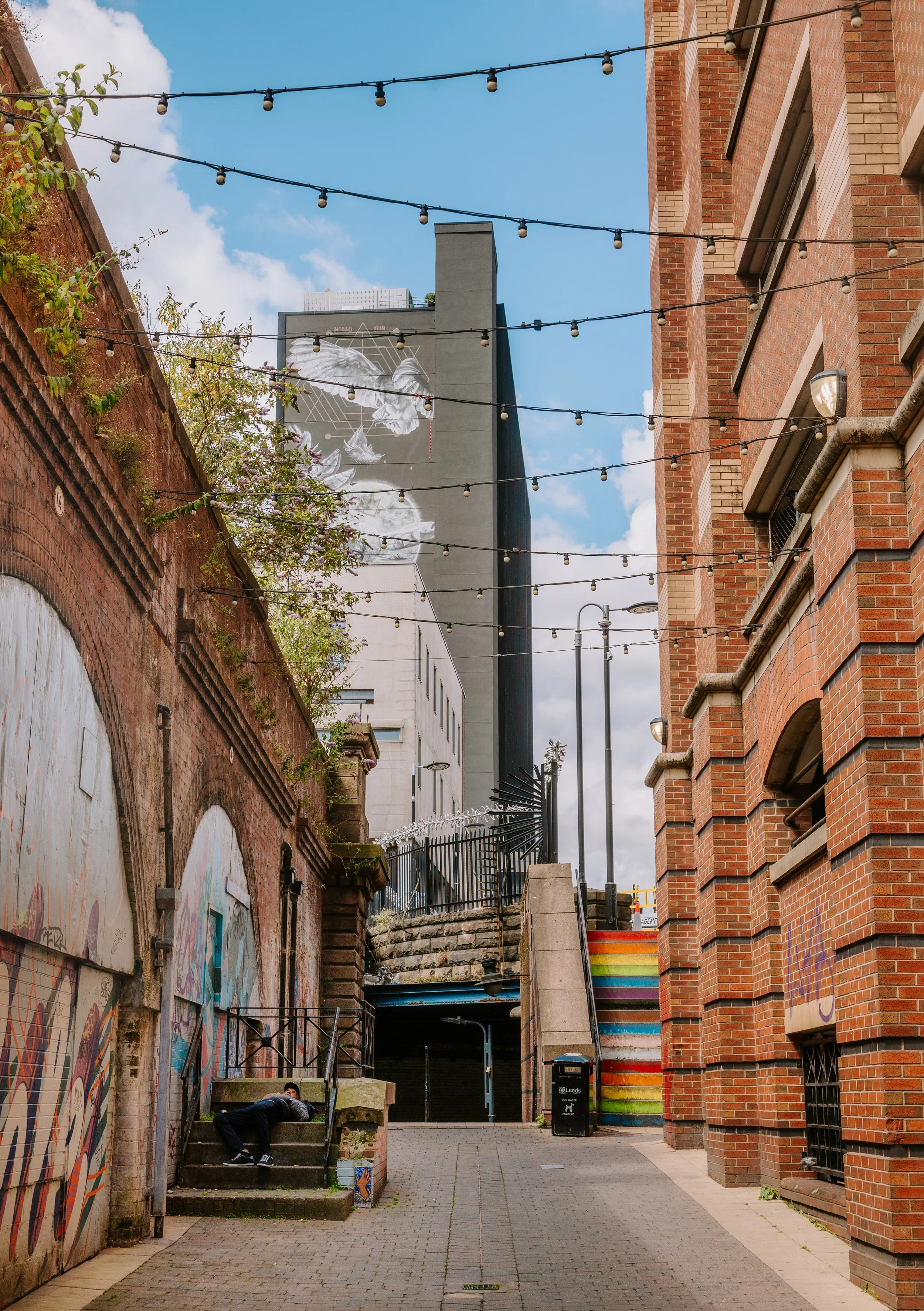 Urban alleyway with brick buildings, graffiti-covered wall on the left, string lights overhead, stairs with graffiti on the left, and a person lying on the stairs. A rainbow-colored staircase and a graffiti mural featuring a dove are visible in the b