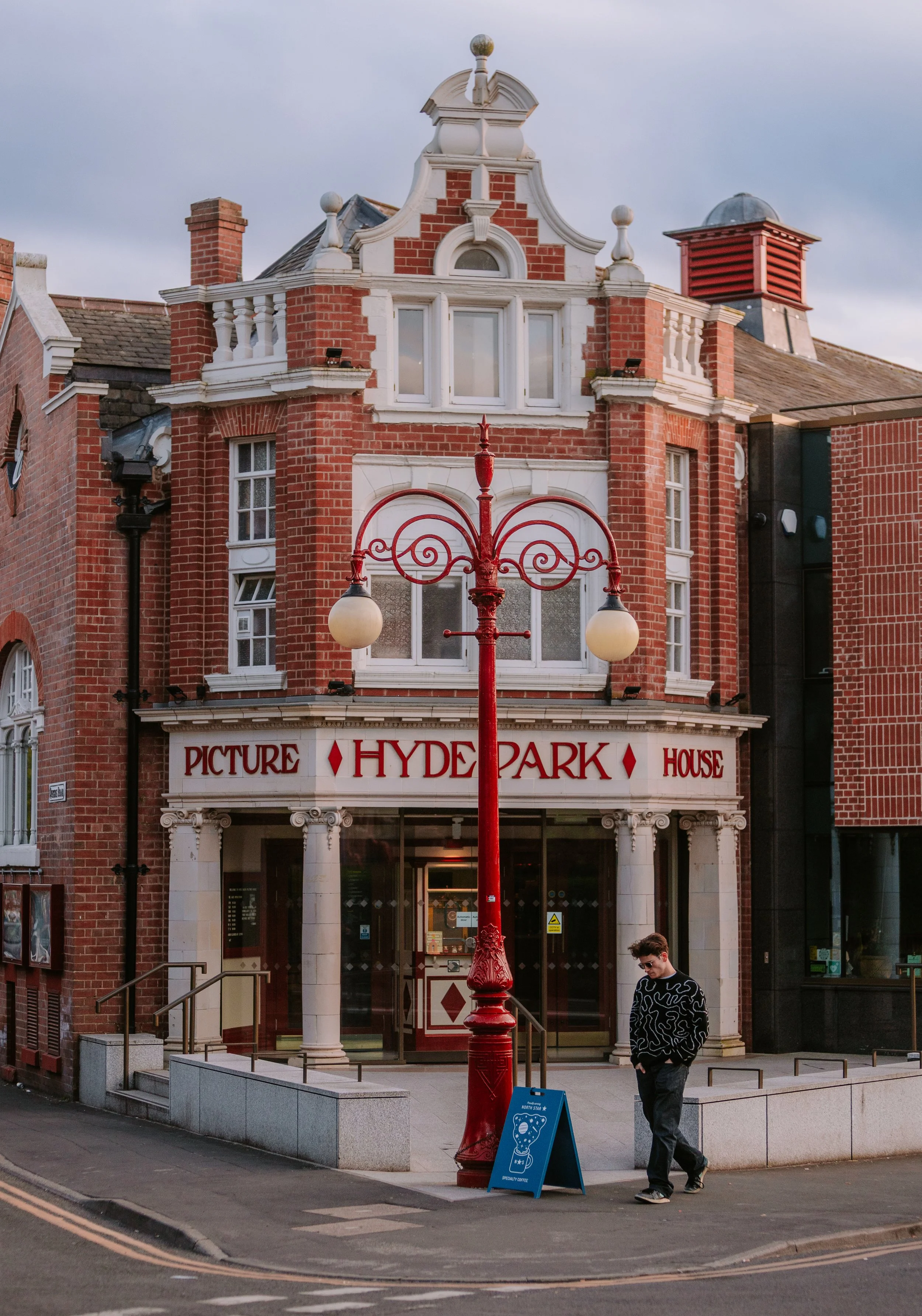 A red brick building with a white sign reading 'Picture Hyde Park House' above the entrance. A red lamppost with two spherical lights is in front. A man with sunglasses and a patterned black and white sweater walks on the side near the building. A bl