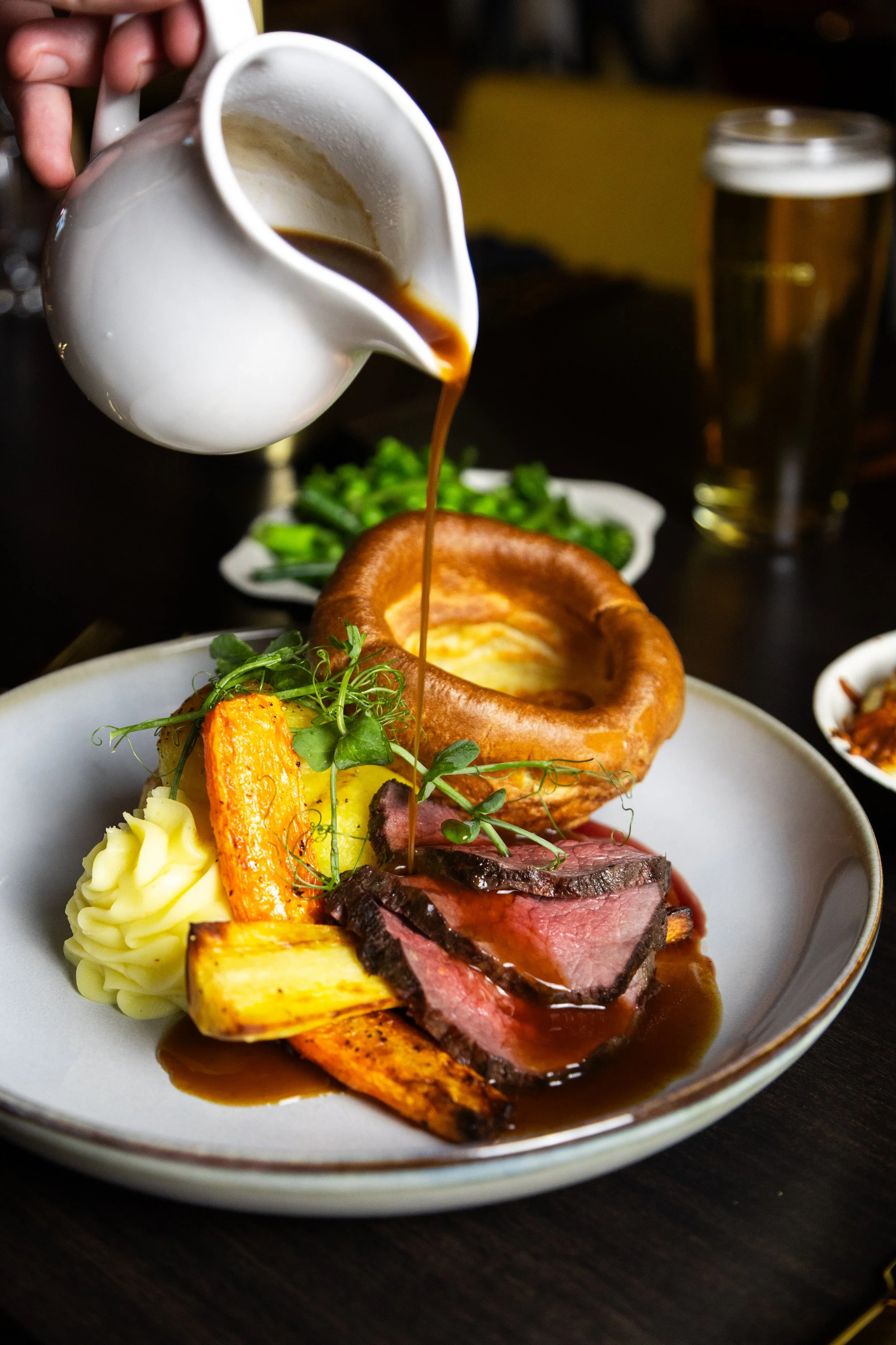 A plate with sliced beef, mashed potatoes, roasted vegetables, and garnished greens, with gravy being poured over the dish. york leeds yorkshire restaurant photographer videographer