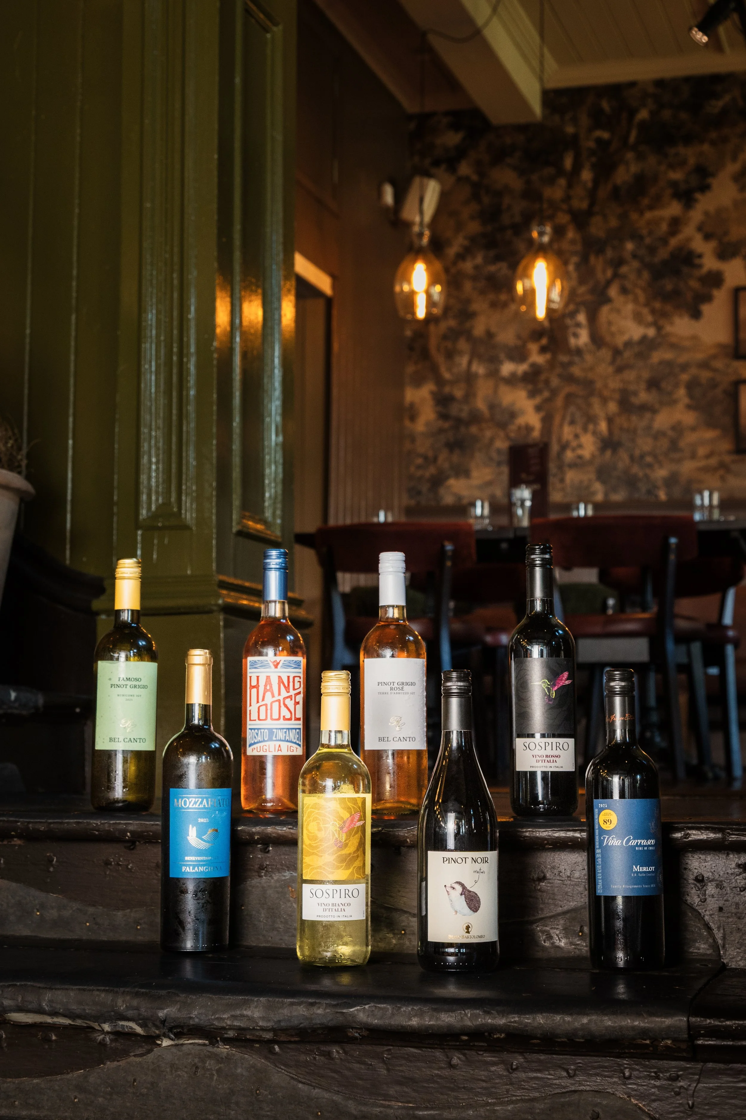 Various bottles of wine on a dark wooden counter in a cozy, dimly lit restaurant with pendant lights and a mural. york leeds yorkshire restaurant photographer videographer
