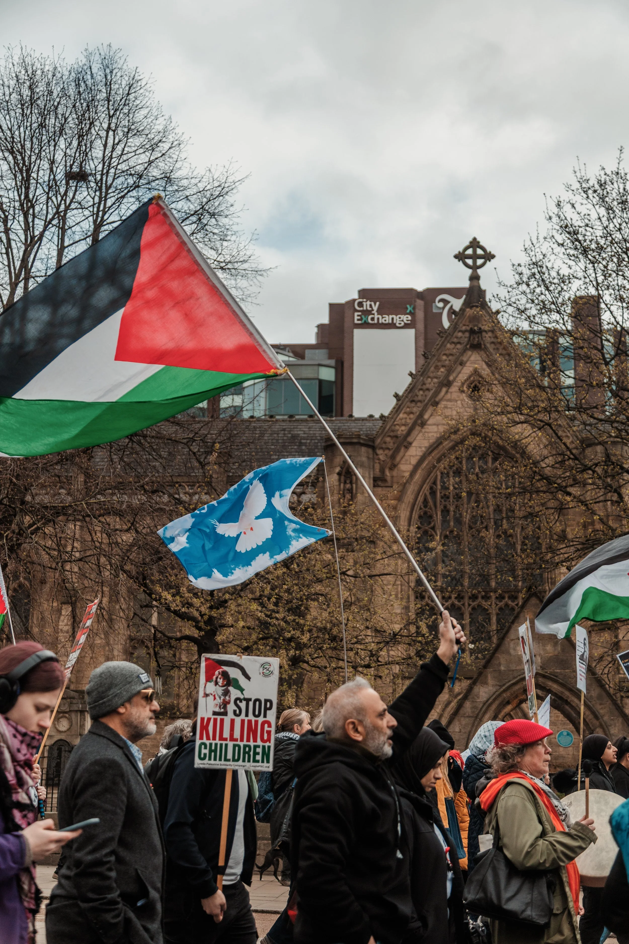 A diverse group of people participating in a protest march, holding signs and flags, with a church and a building labeled City Exchange in the background.