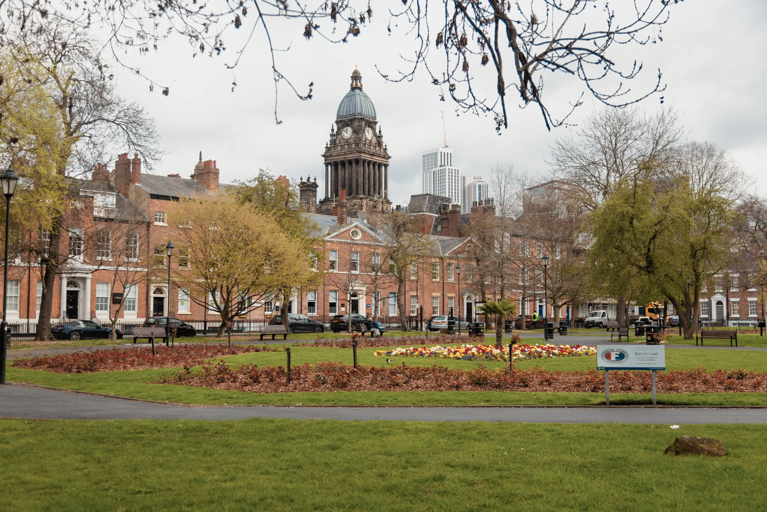 A city park with a green lawn, benches, trees, and colorful flower beds in the foreground. In the background, historic brick buildings and a prominent clock tower with a dome and clock faces. Modern high-rise buildings are visible behind the clock to