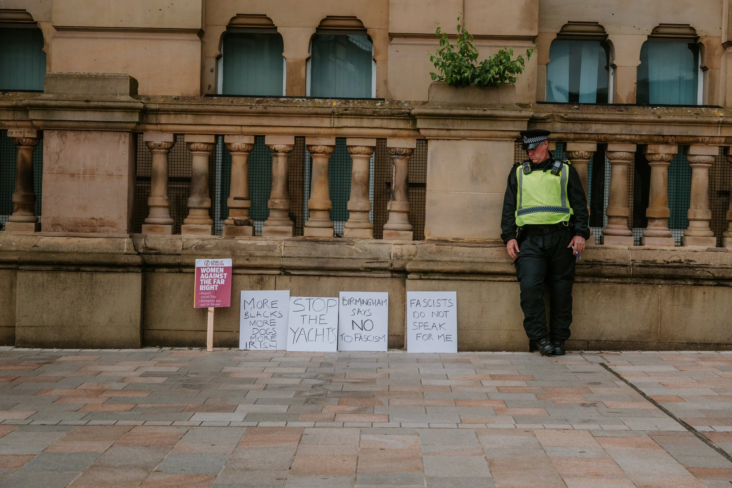 A police officer standing next to a series of protest signs on the ground, in front of a historical building with stone columns and small windows. The signs contain handwritten messages opposing fascism, supporting refugees, and criticizing racist an