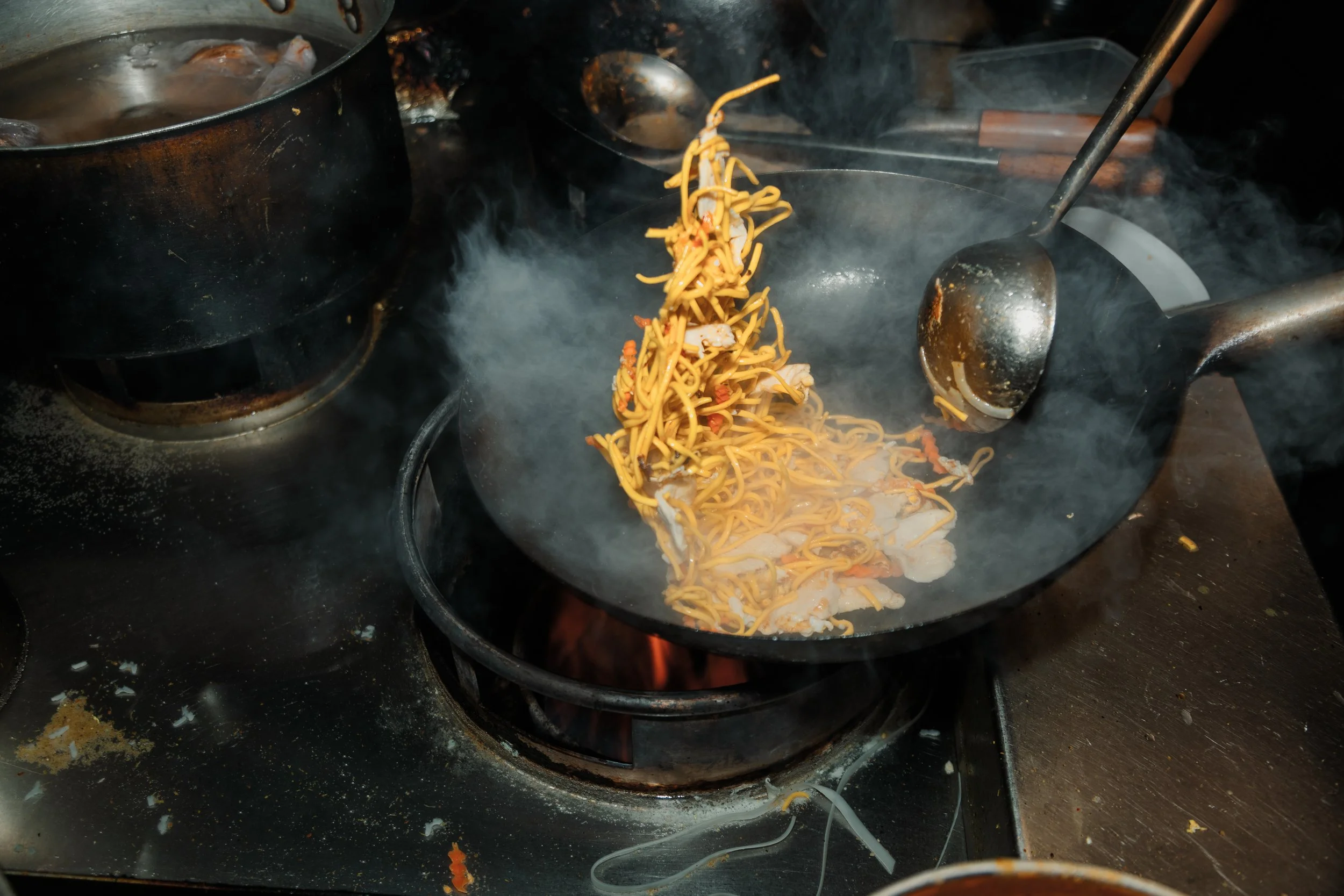 Stir-frying noodles and vegetables in a black wok on a stove, with steam rising. york leeds yorkshire restaurant photographer videographer