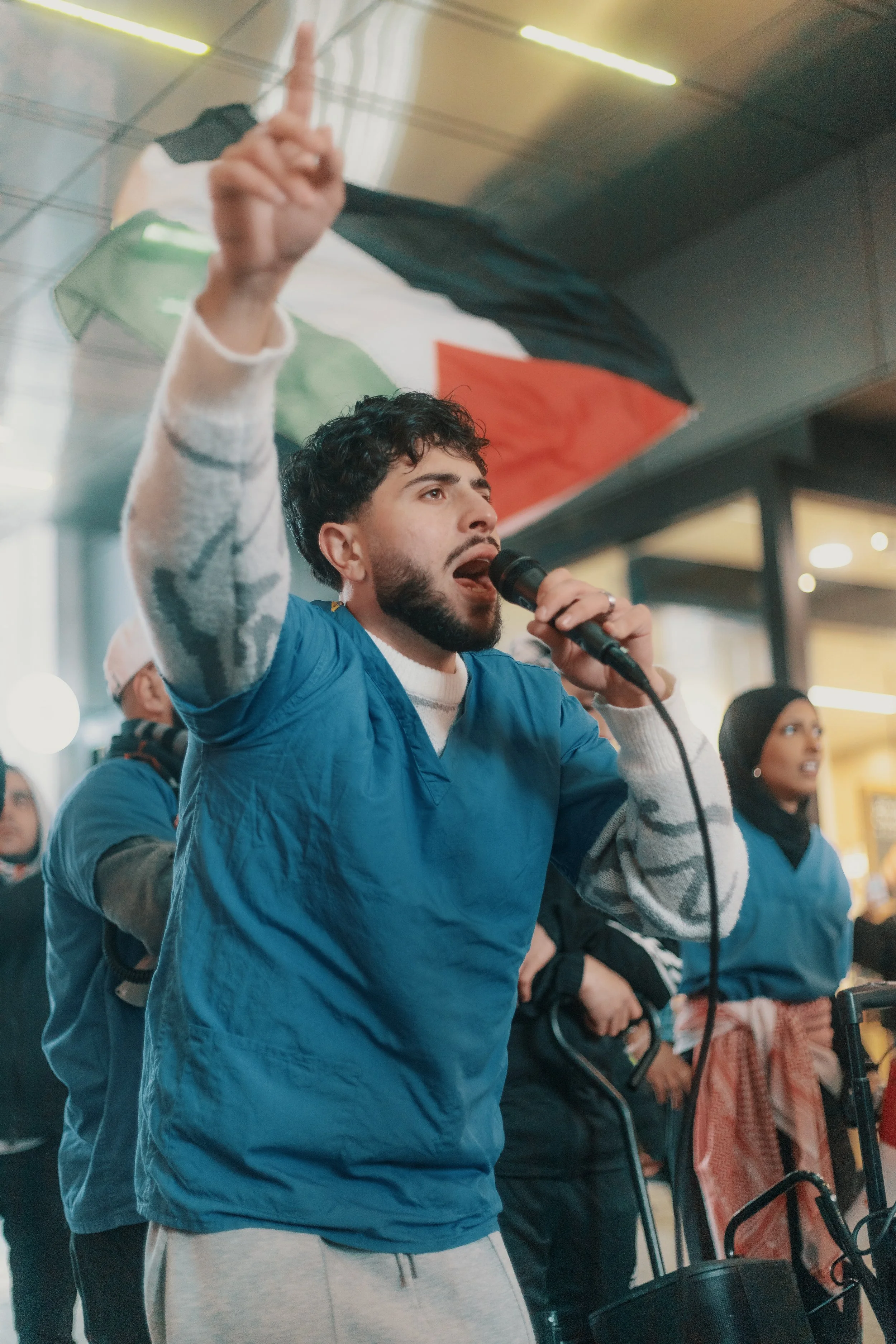A man with black curly hair and a beard, wearing a blue vest and gray sweatpants, holding a microphone with his mouth open and his hand raised, making a peace sign gesture. In the background, a woman wearing a black hijab and a blue vest, with her ha