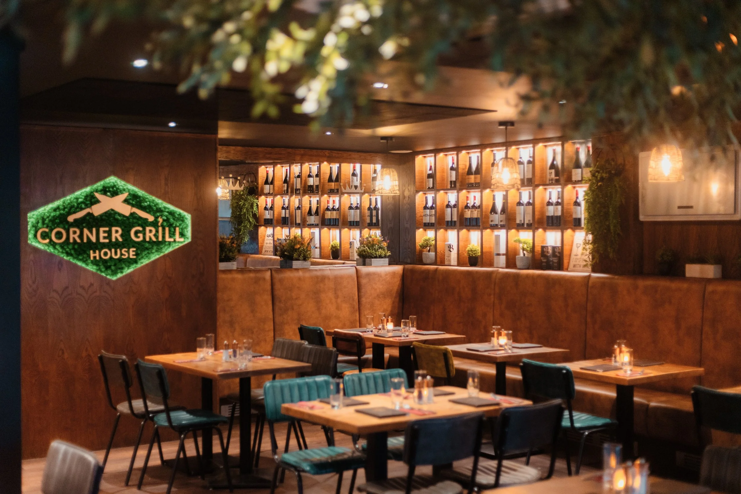 Interior of a restaurant with wooden tables set with glasses and candles, and a wall behind filled with shelves of wine bottles, decorated with hanging lights and greenery. york leeds yorkshire restaurant photographer videographer