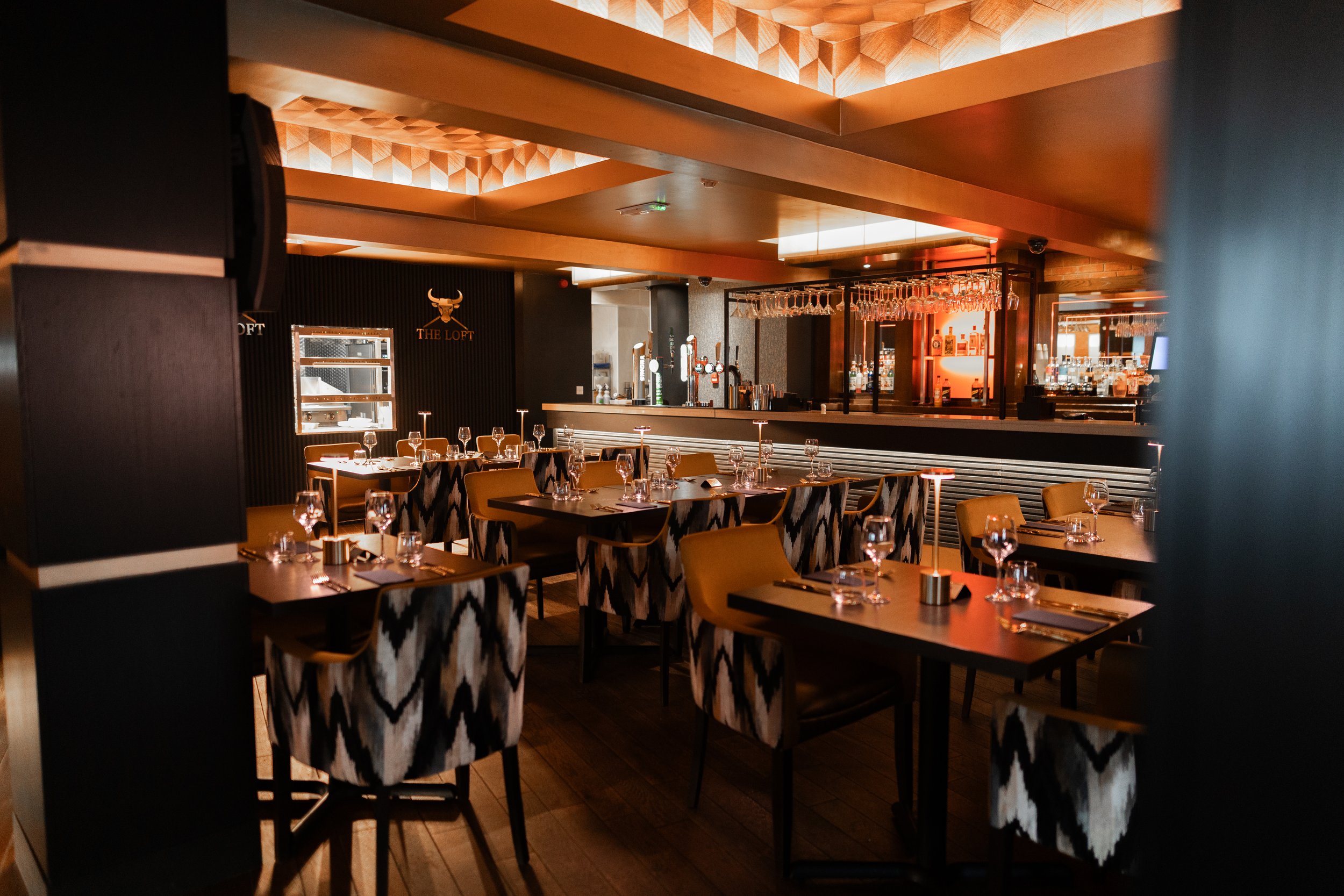 A modern restaurant interior with tables set with wine glasses and silverware, patterned chairs, and warm lighting. The bar area with hanging glasses and bottles is visible in the background, along with a logo that says 'The Loft' with a bull's head.