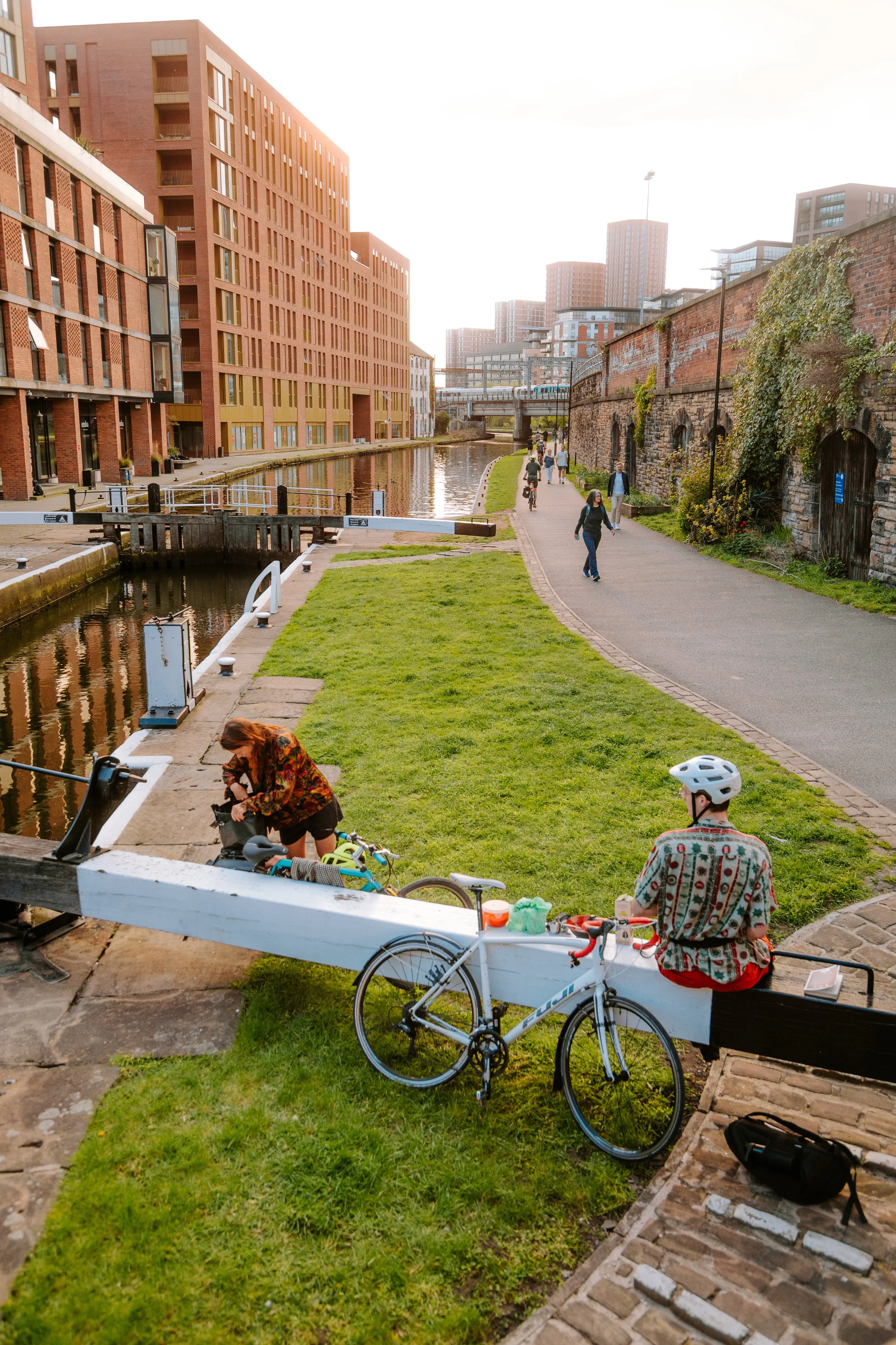 People walking, biking, and sitting near leeds canal and train station and mustard wharf with modern buildings on one side and an old brick wall on the other, during daytime.