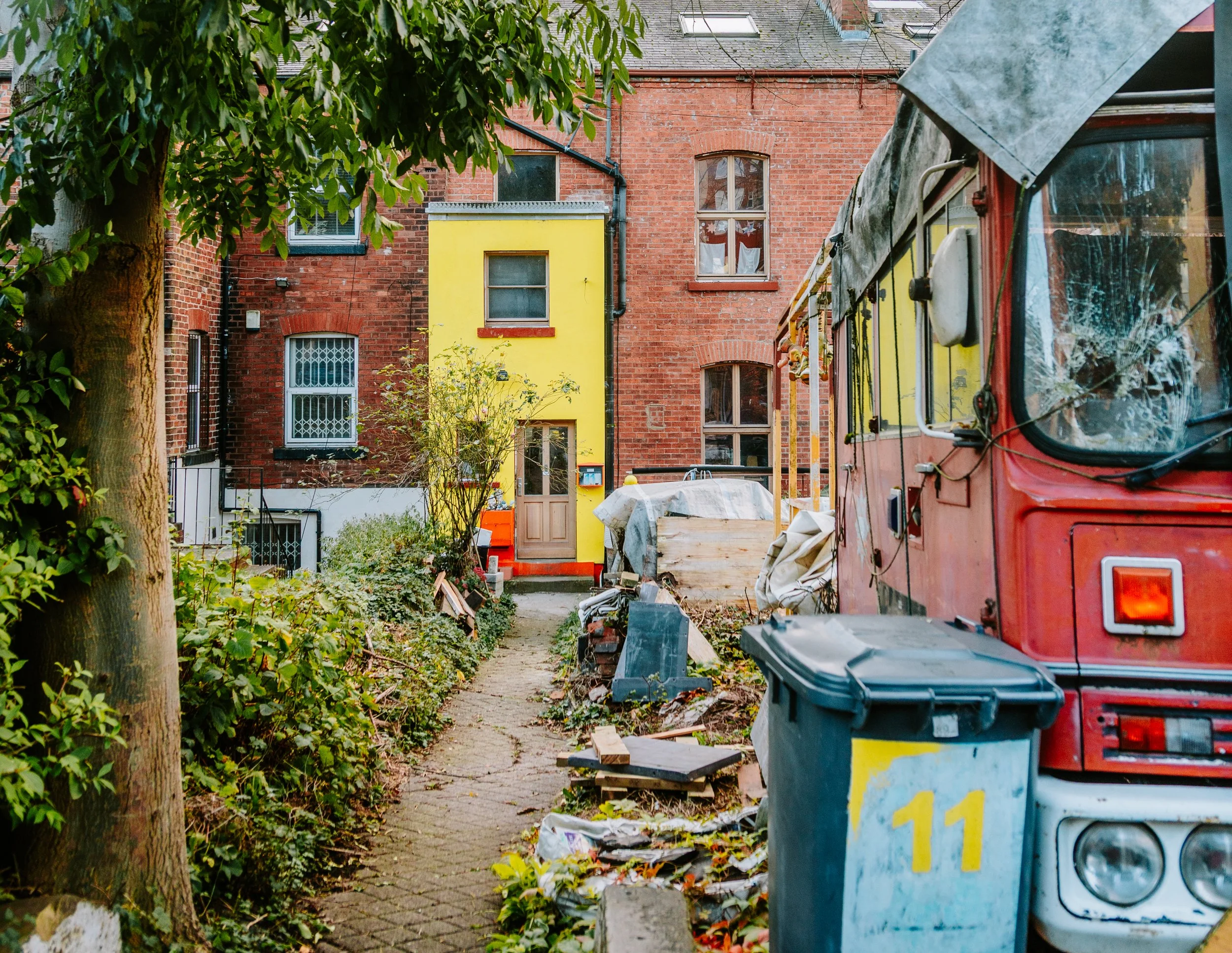 Rear view of a narrow alleyway with trash bins, debris, and a construction area in front of red brick and yellow buildings, with a tree partially obscuring the view on the left. hyde park leeds.