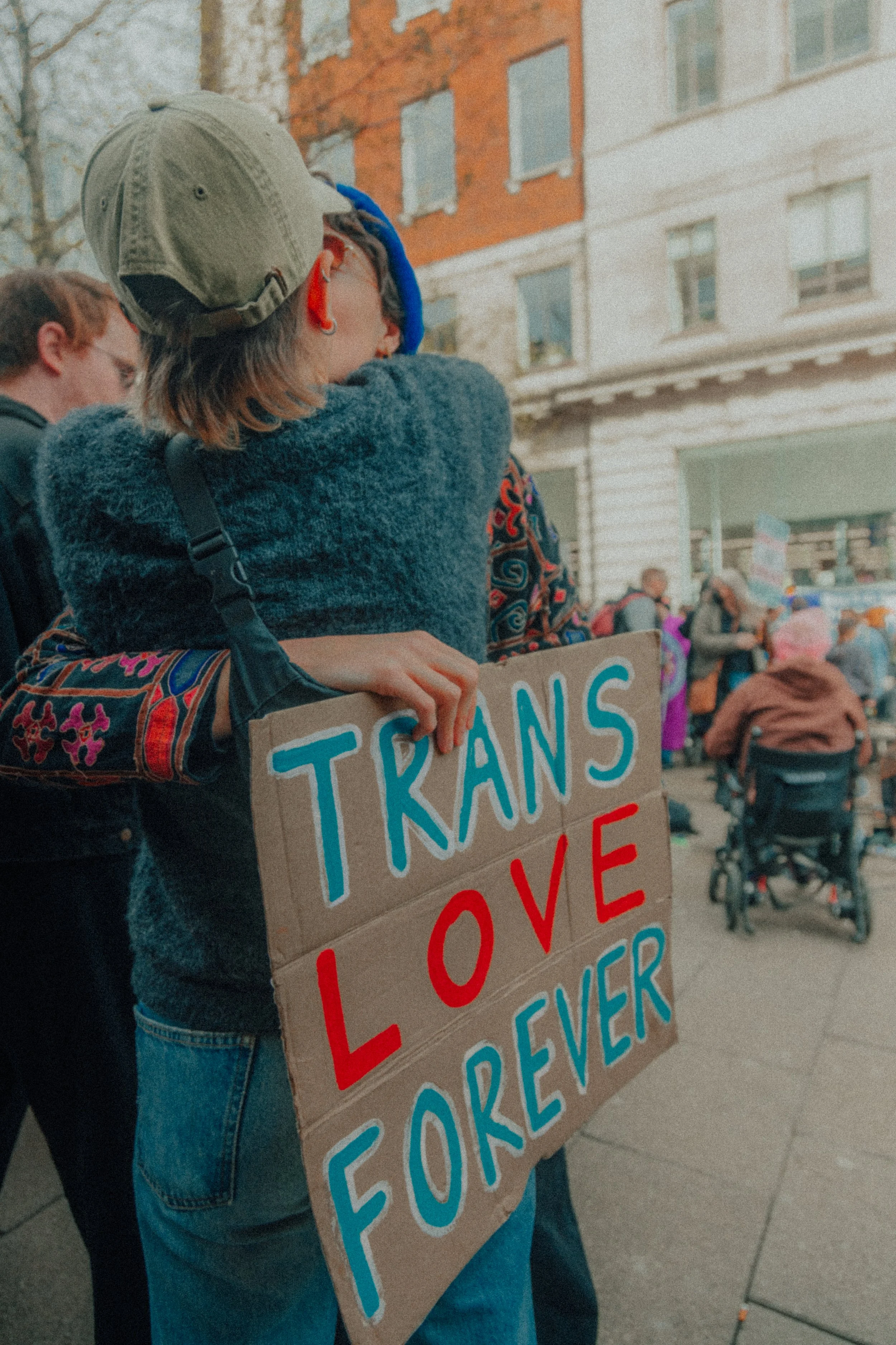A person holding a cardboard sign that says 'Trans Love Forever' in a crowd. The person is wearing a beige cap, large glasses, earrings, a blue beret, and a fuzzy coat. Other protesters and buildings are visible in the background.