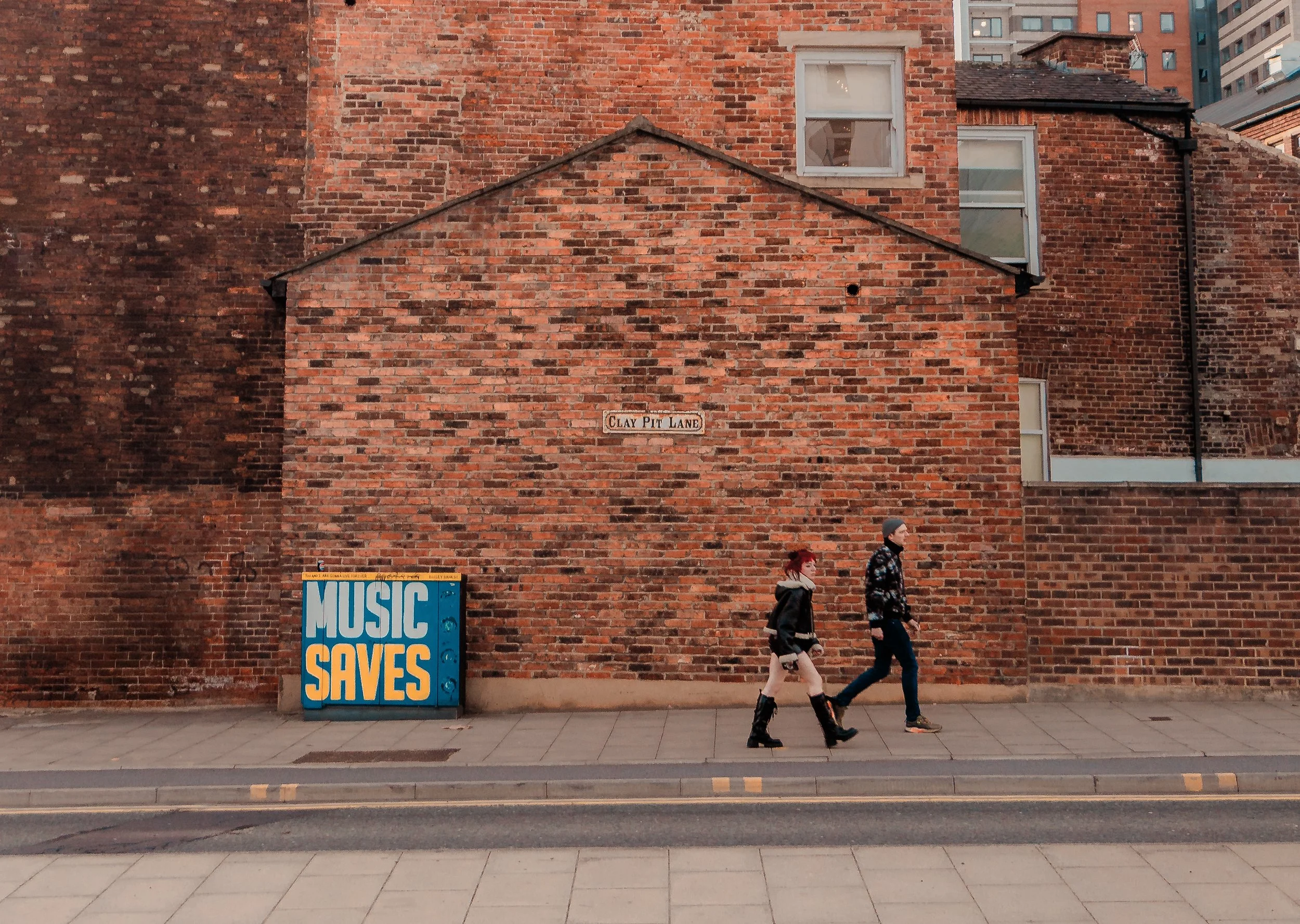 A city sidewalk scene with two pedestrians walking past a large red brick building. The building has a small sign that reads 'Clay Pit Lane' and a colorful sign that says 'Music Saves' next to it. Part of a higher building with numerous windows and a