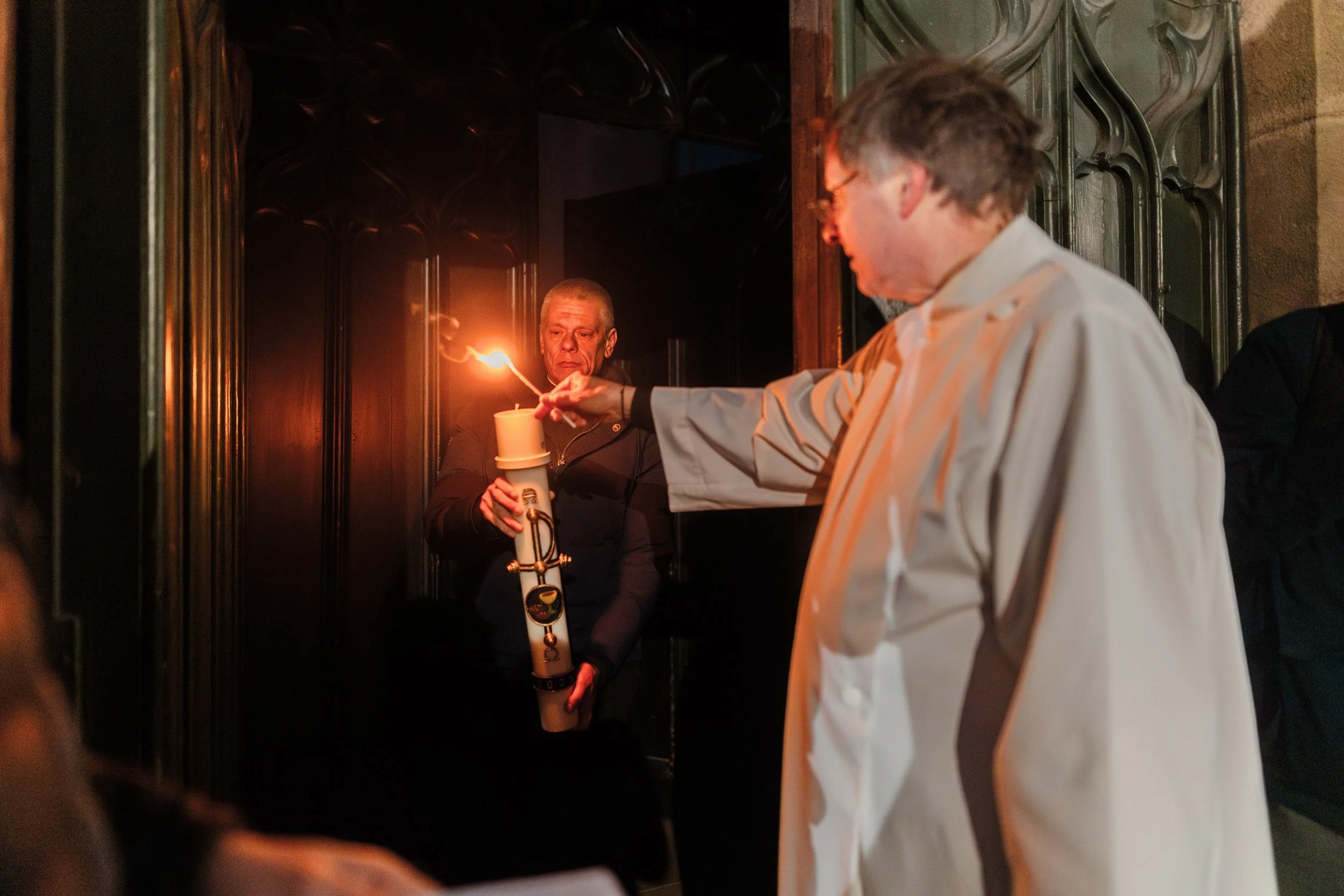 A man holding a lit candle and a stick with a torch, standing near a young person in a white coat inside a dark, ornate room.