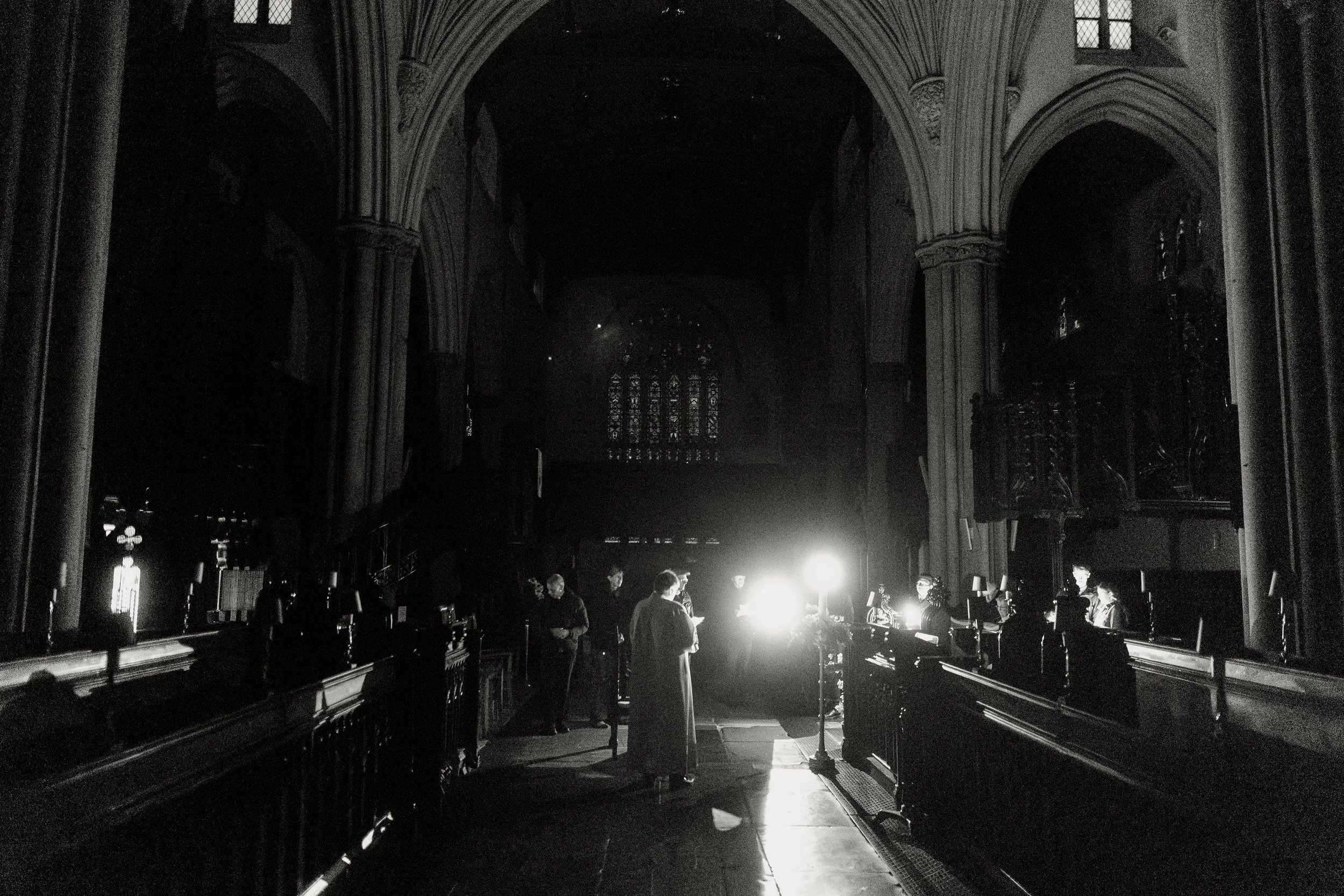 Interior of a Gothic-style church with pointed arches and stained glass windows. People are gathered around a table with bright light, creating silhouettes against the dark interior.