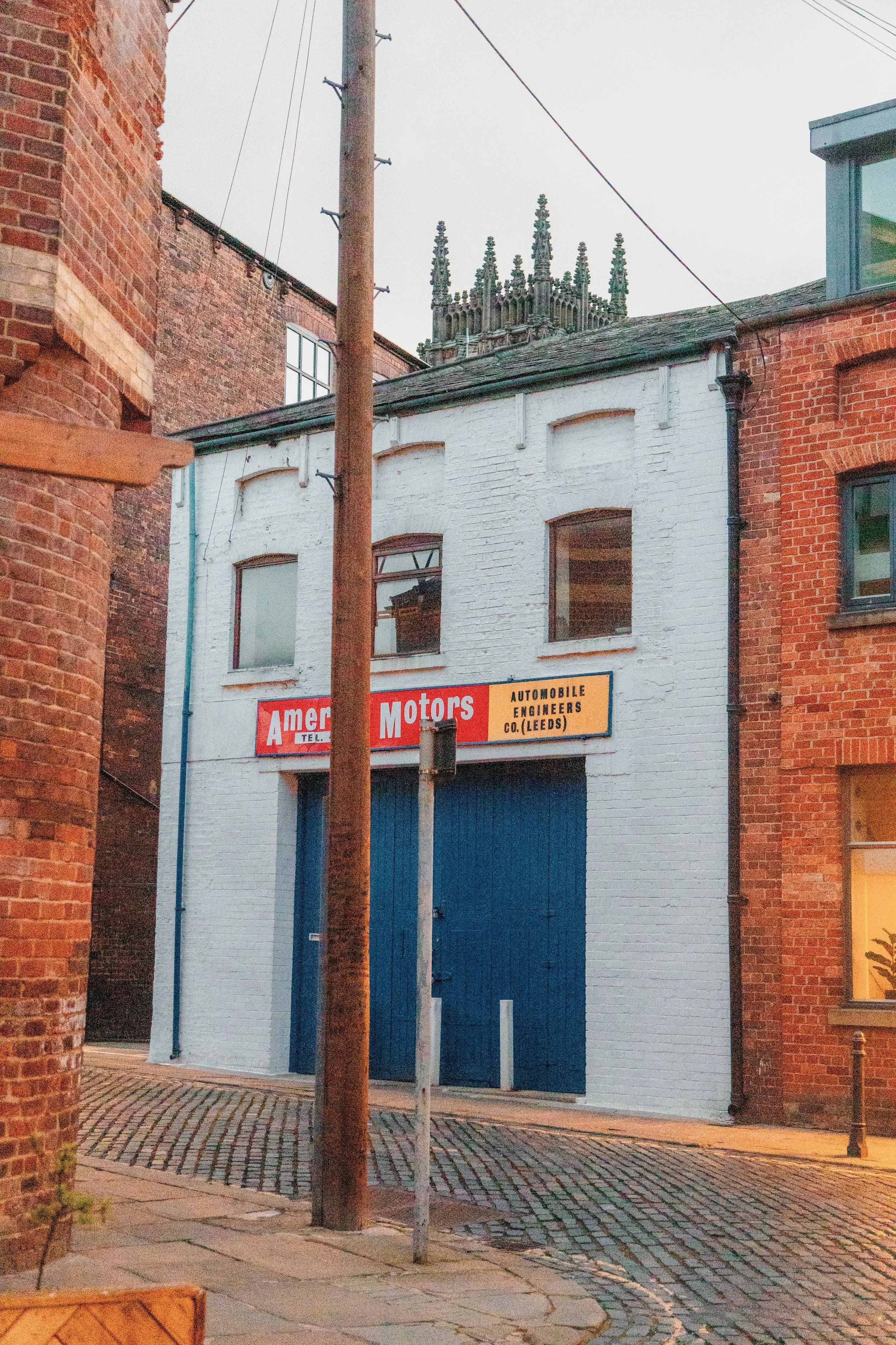 A street view showing a brick building on the left, another red brick building on the right, a white building with blue doors in the middle, a wooden utility pole, and a church steeple in the background. There is a sign for 'Amer Motors' on the white