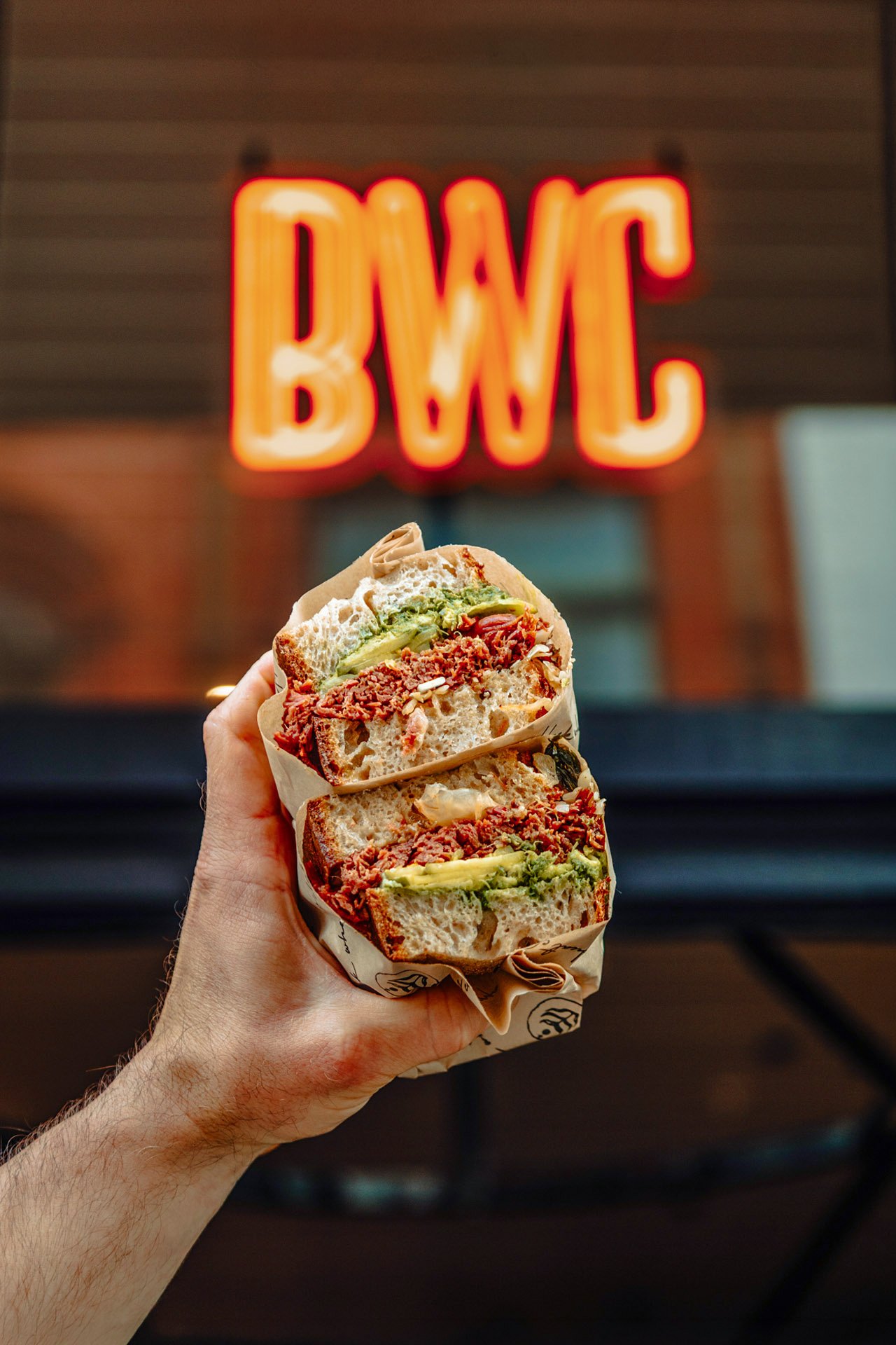 A hand holding a large sandwich with various ingredients including shredded meat, lettuce, and other fillings in front of a lit neon sign that reads 'BWC' on a wooden background. york leeds yorkshire restaurant photographer videographer