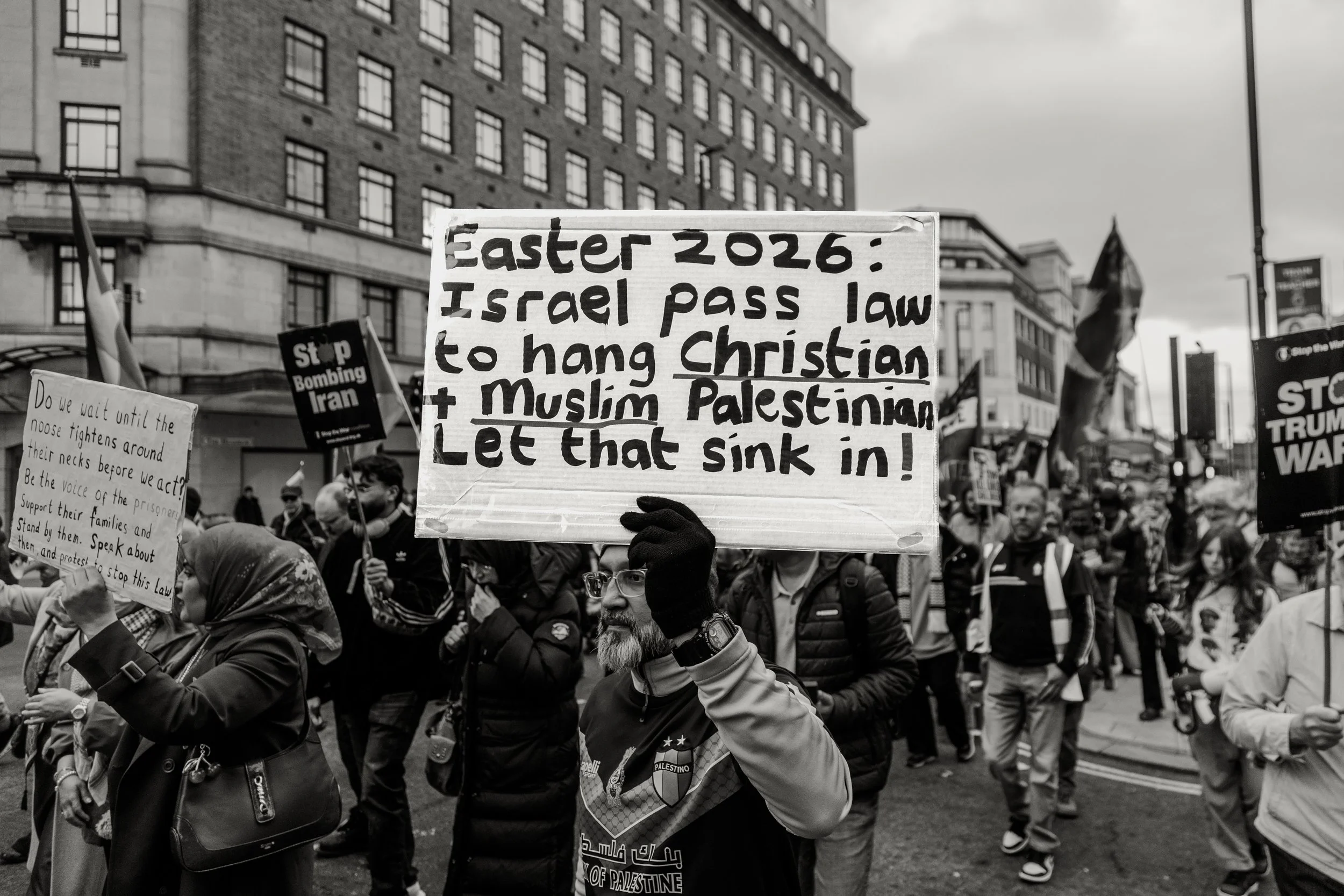 A crowd of protesters in an urban setting holding various signs, one prominently displays a message about Israel's 2026 law and Palestinian issues, with other signs visible. The scene appears to be during a demonstration or rally.