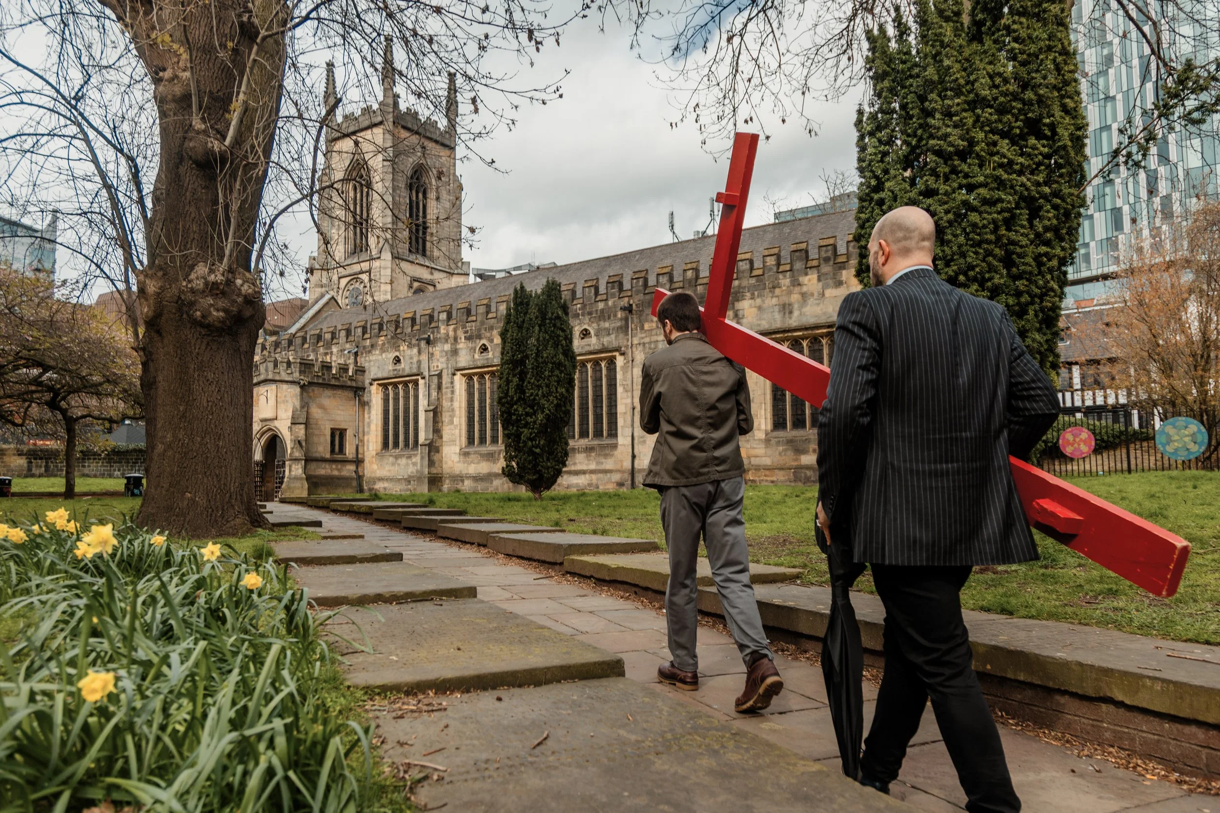 Two men in suits walking up stone steps in a park with a church and modern glass building in the background. One man carries a large red wooden cross. The park has a tree with bare branches and yellow flowers in the foreground.