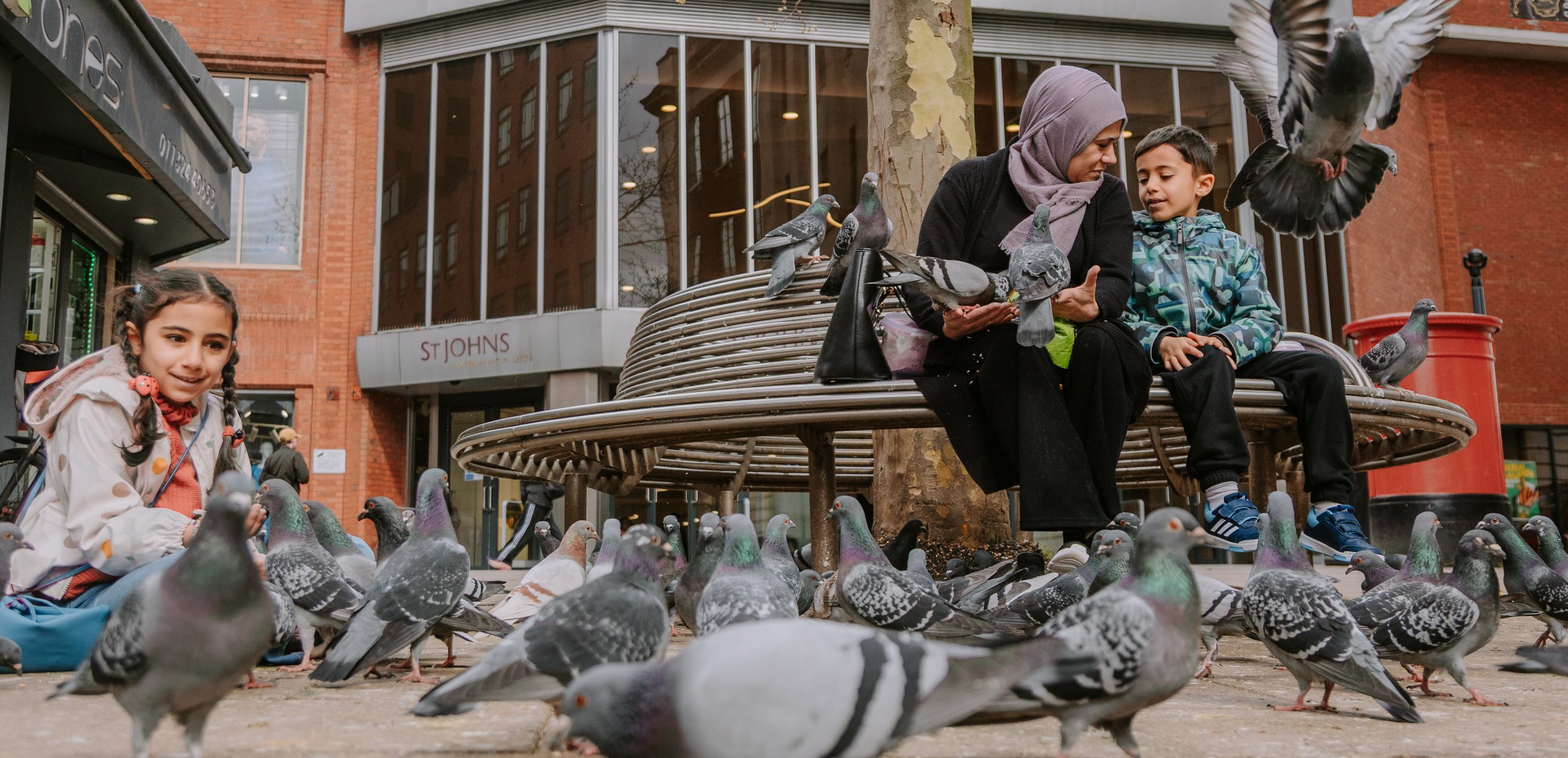 A woman and a boy are sitting on a park bench surrounded by pigeons on the ground and in the air, with a girl nearby also with pigeons, in an urban area with buildings and a store sign reading 'St. Johns' in the background.