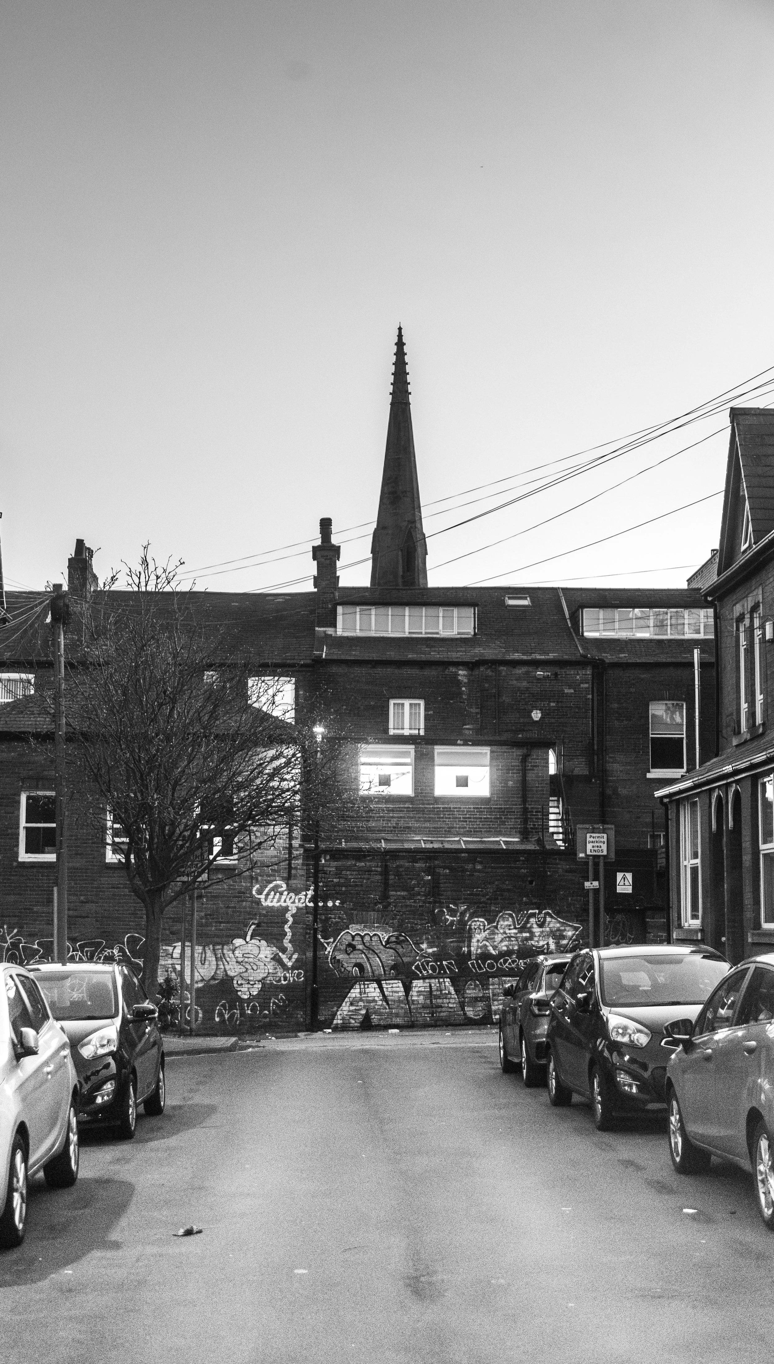 Black and white photo of a street with parked cars, a graffiti-covered wall, and a building with a church steeple in the background.
