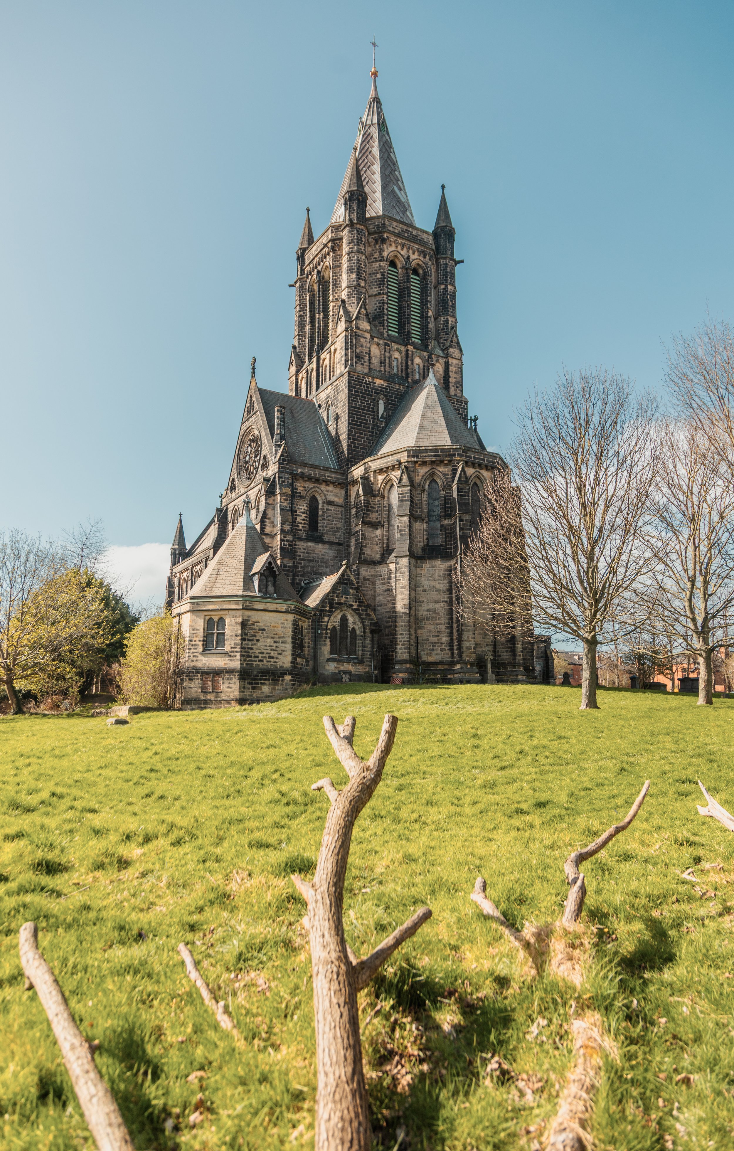 A tall, old stone church with pointed steeples and large arched windows, set against a clear blue sky and surrounded by green grass and trees.