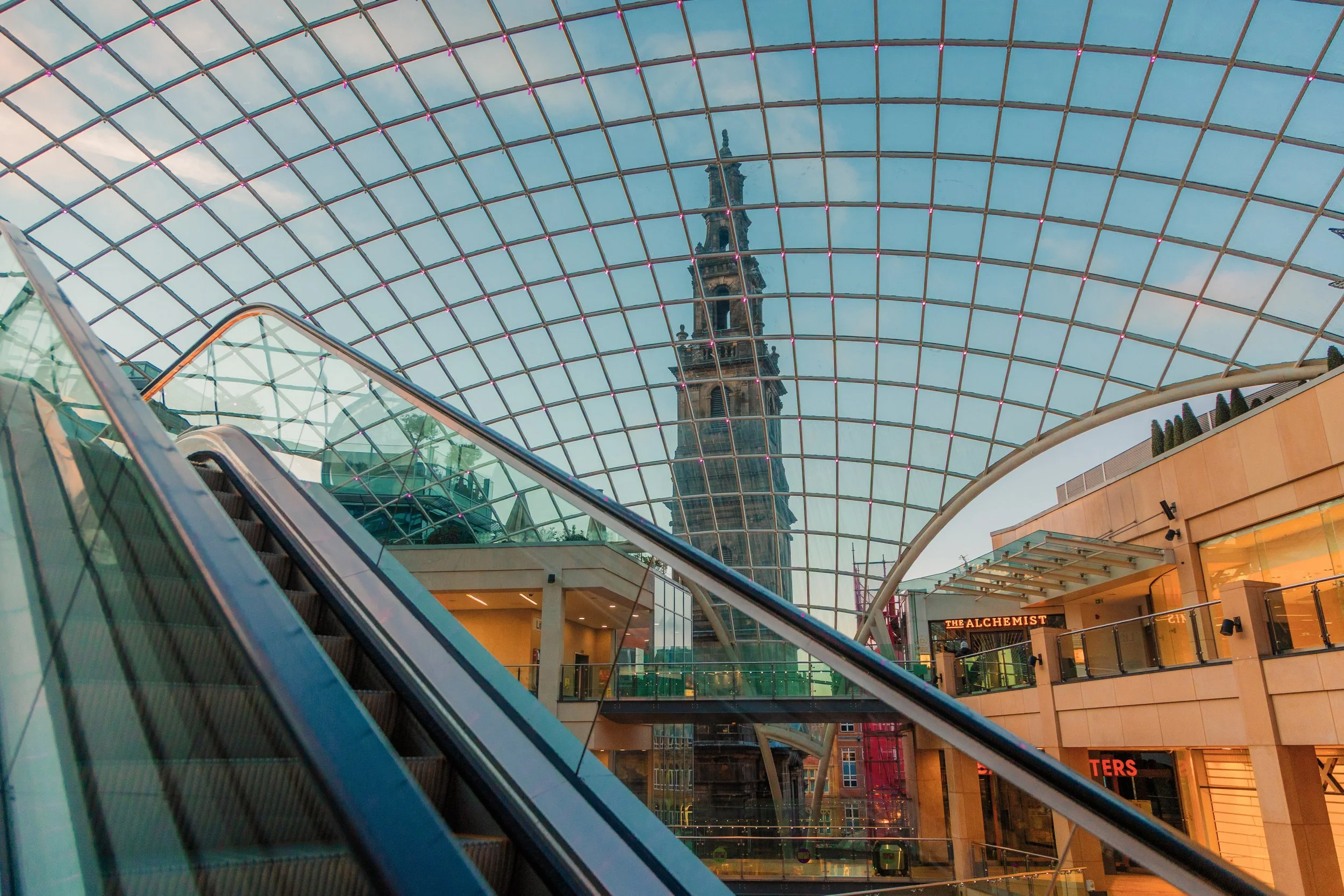 A view through a glass roof of a shopping mall, showing an escalator and a tall historic clock tower in the background. Leed trinity shopping centre.