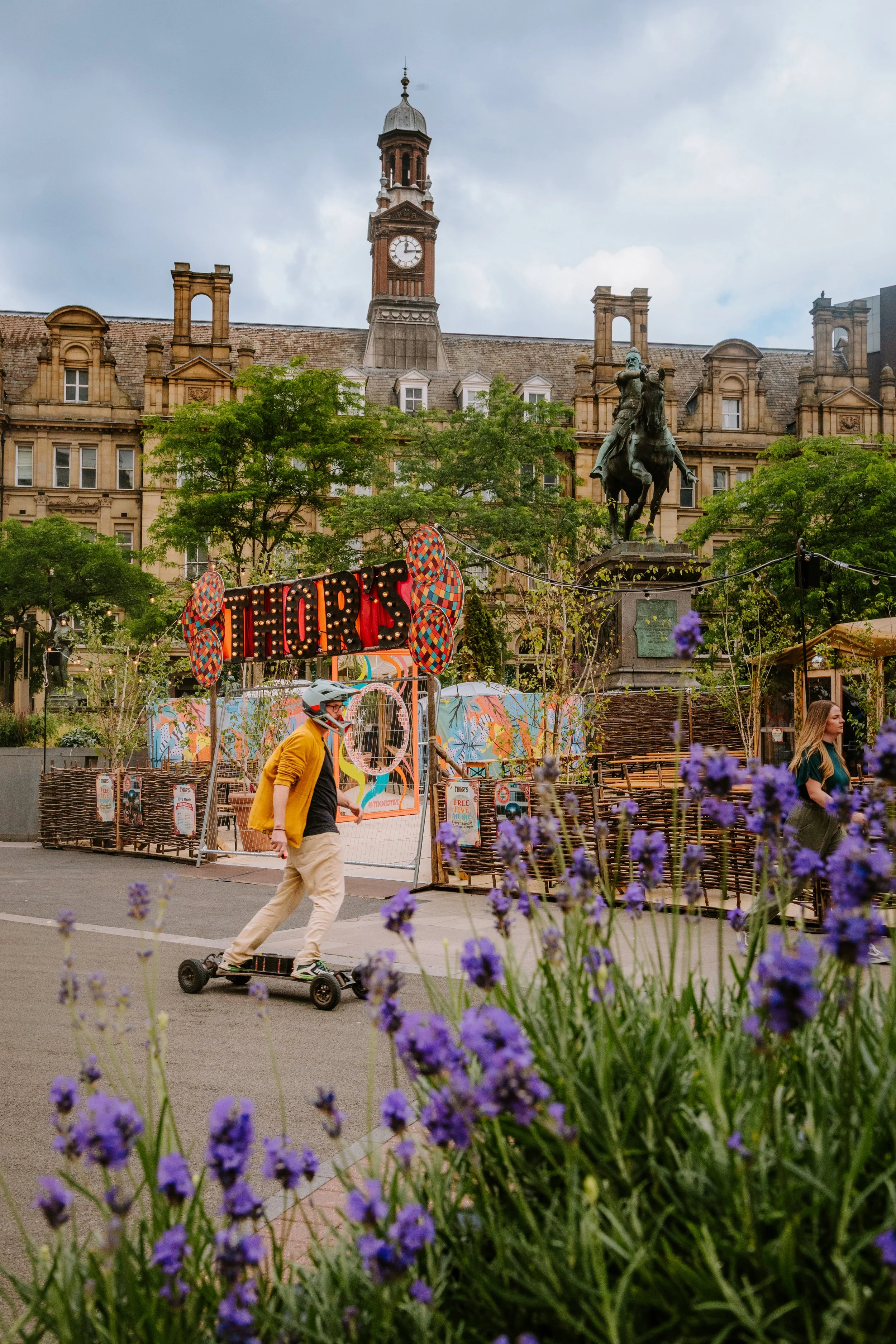 A city square with a historic building featuring a clock tower in the background, a statue on horseback, and a street fair or festival with purple flowers in the foreground. boosted board, skateboard hoverboard.