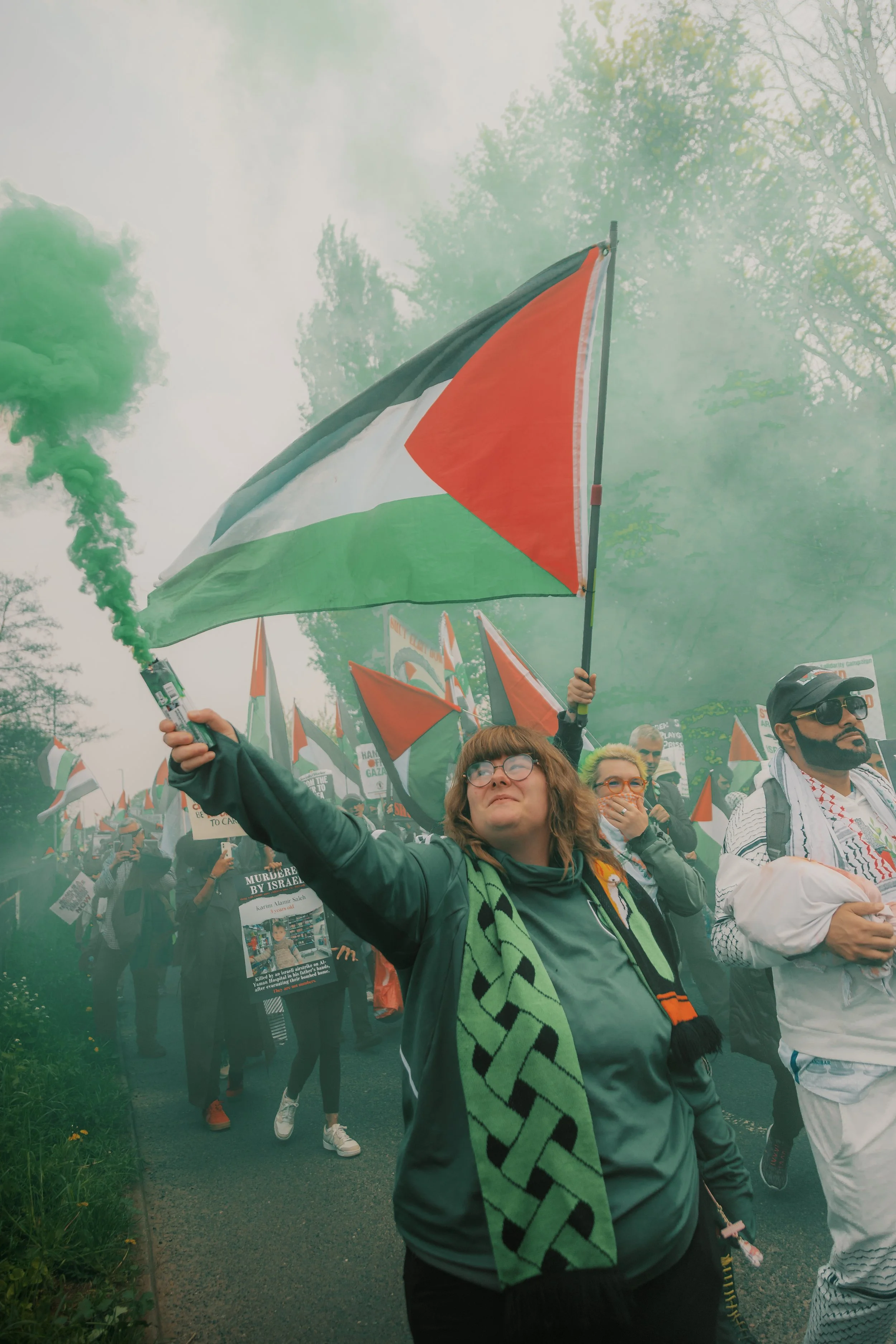 People participating in a protest or march holding flags, with some wearing scarves and masks, and one person in front holding a green smoke flare.