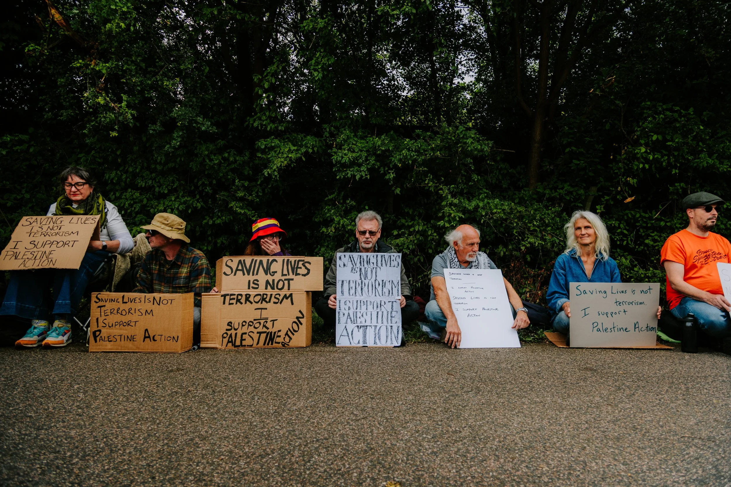 Group of people sitting on the ground holding protest signs supporting Palestine and opposing terrorism in a park with trees in the background.