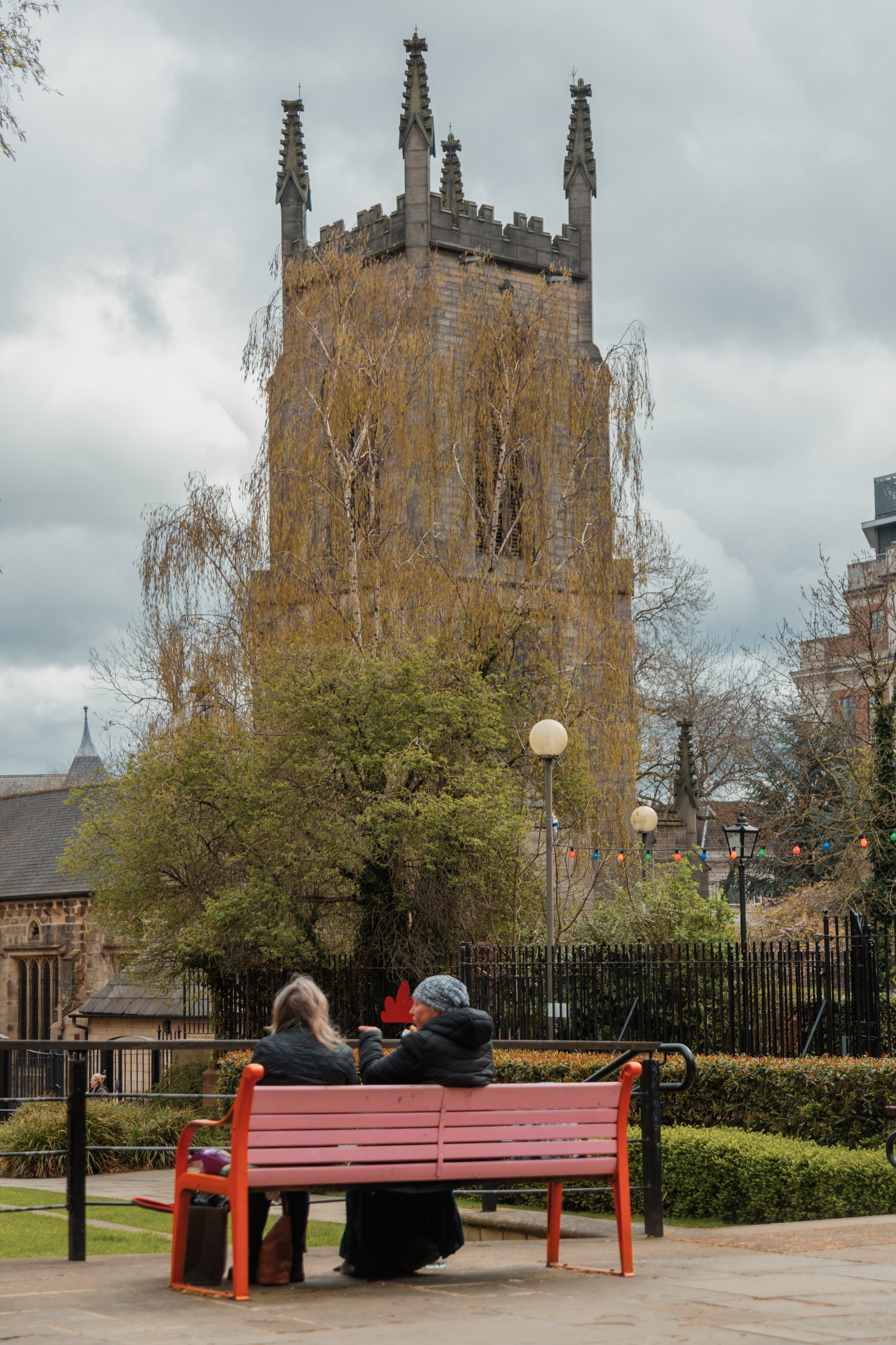 A park with pink benches, lush green bushes, and two women sitting on one of the benches. In the background, there is a tall, gothic-style church tower with four spires and decorative elements, partially covered by leafless trees. Overhead, the sky i