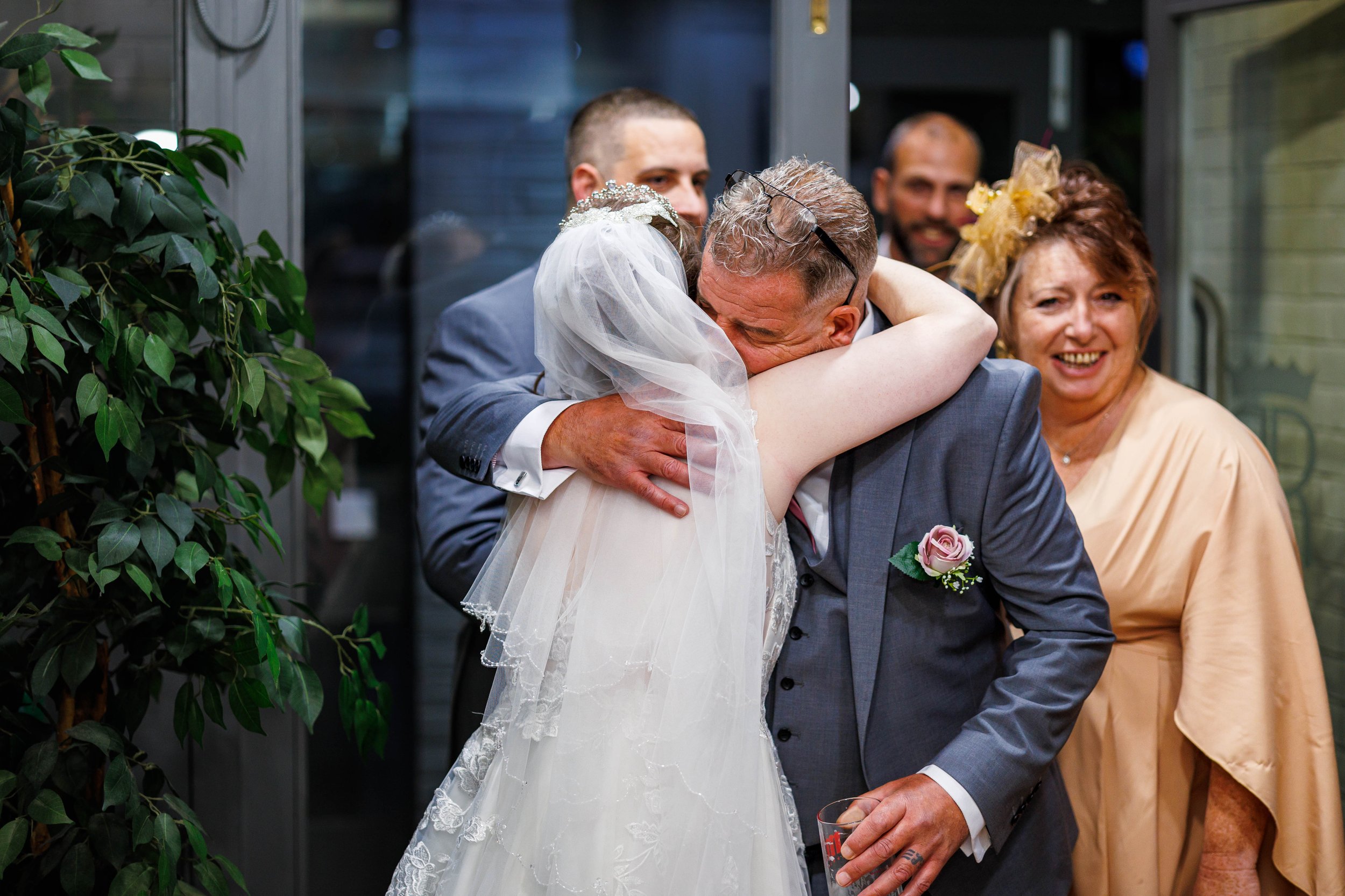 Bride hugging a man at a wedding reception with smiling guests in the background.