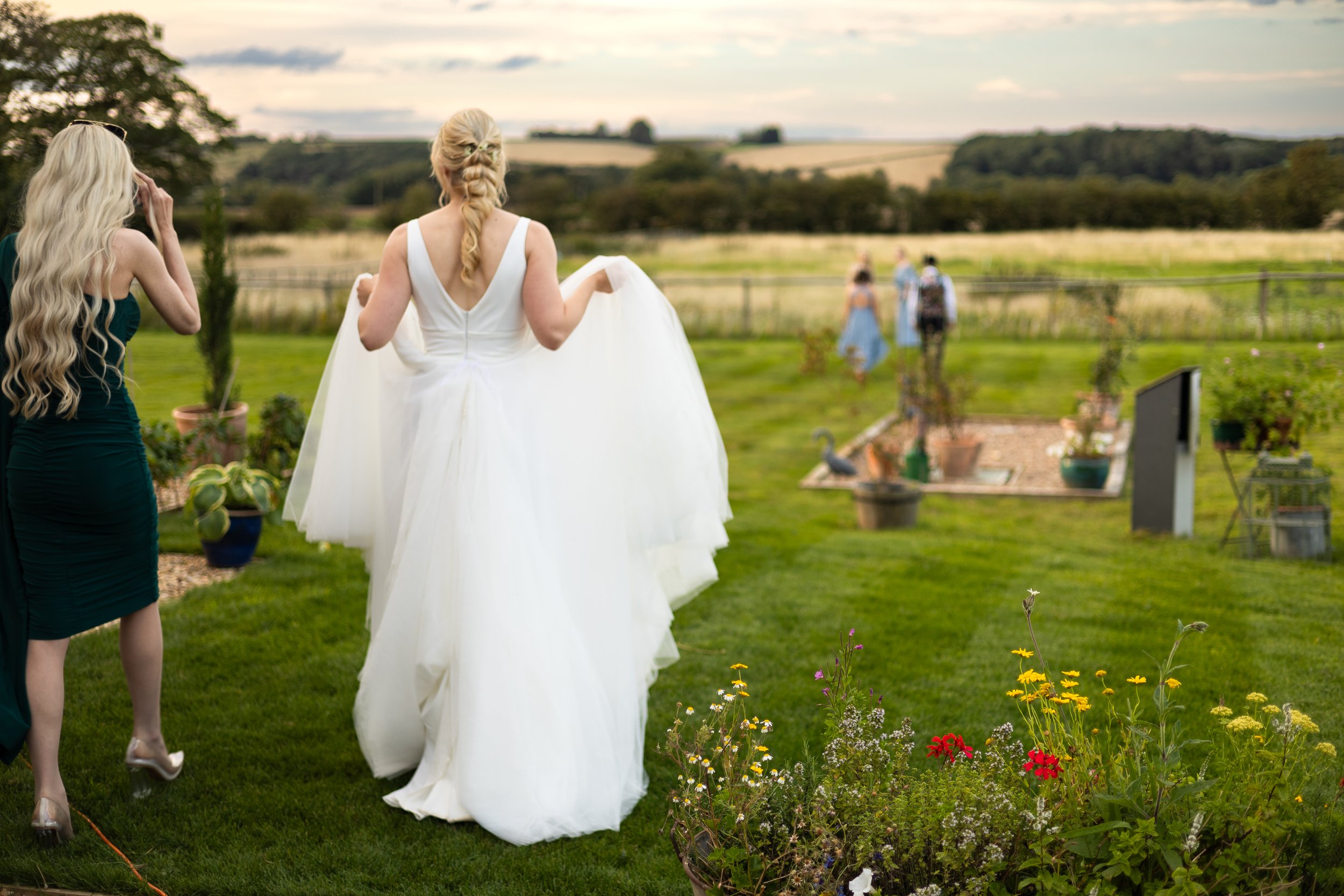 Bride in white dress walking in garden with floral and countryside scenery at outdoor event.