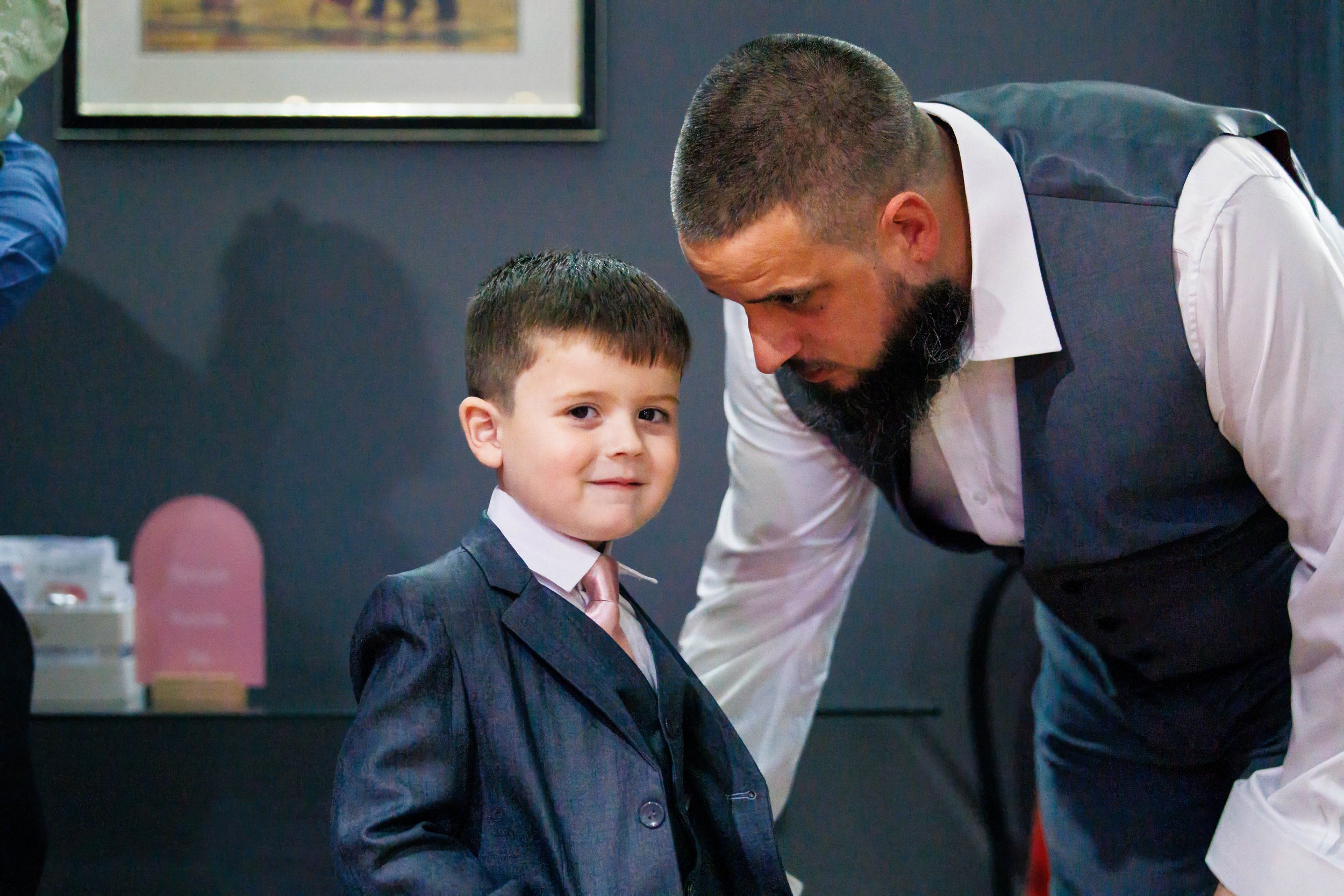 A young boy in a suit and tie stands next to a bearded man in a white shirt and vest, who is leaning in to speak with him.