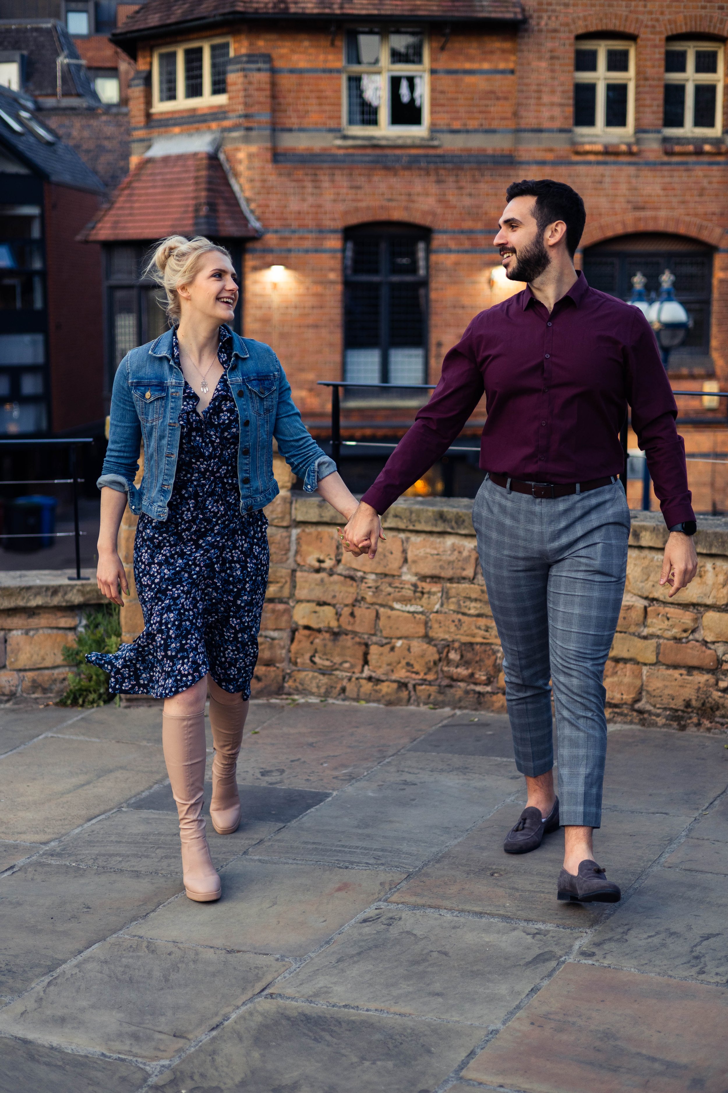 A smiling couple holding hands while walking, the woman wearing a blue floral dress and denim jacket, the man in a maroon shirt and plaid pants; urban brick building in background.